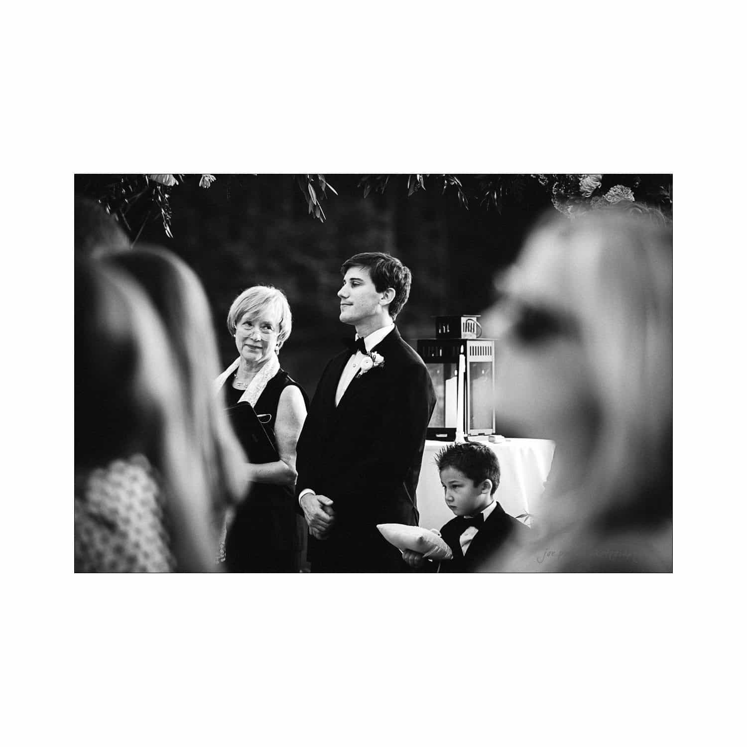 A black and white photo of a formal gathering. A man in a tuxedo stands beside an older woman, both looking ahead. A young boy in a suit kneels beside them. Blurred figures are visible in the foreground.