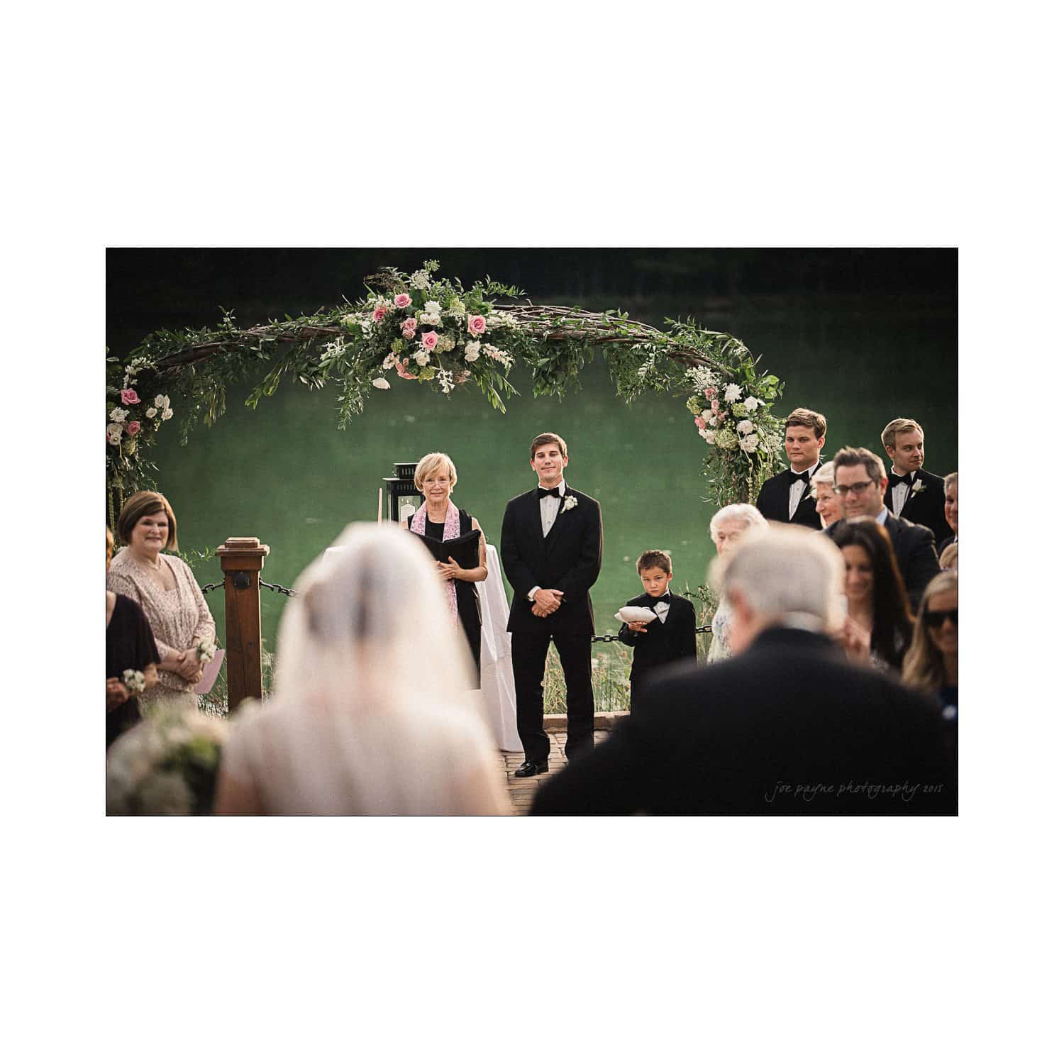 A wedding ceremony by a lake, featuring a floral archway. The groom stands with a young boy beside him, awaiting the bride, who is seen from behind. Guests are seated, and an officiant is present.