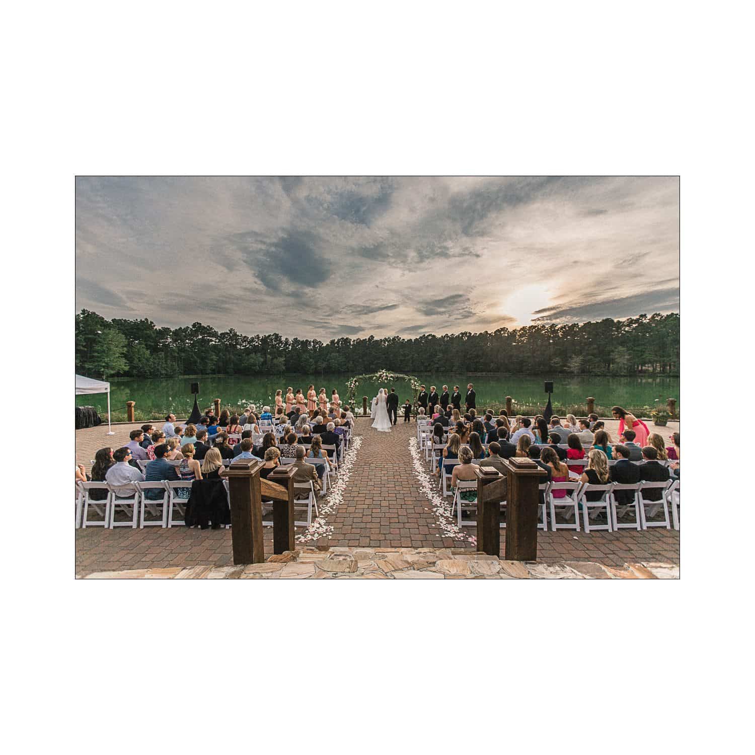 A wedding ceremony by a lake at sunset. The bride and groom stand under an arch, with the wedding party on each side. Guests are seated in rows, facing the couple. Trees and a cloudy sky are in the background.