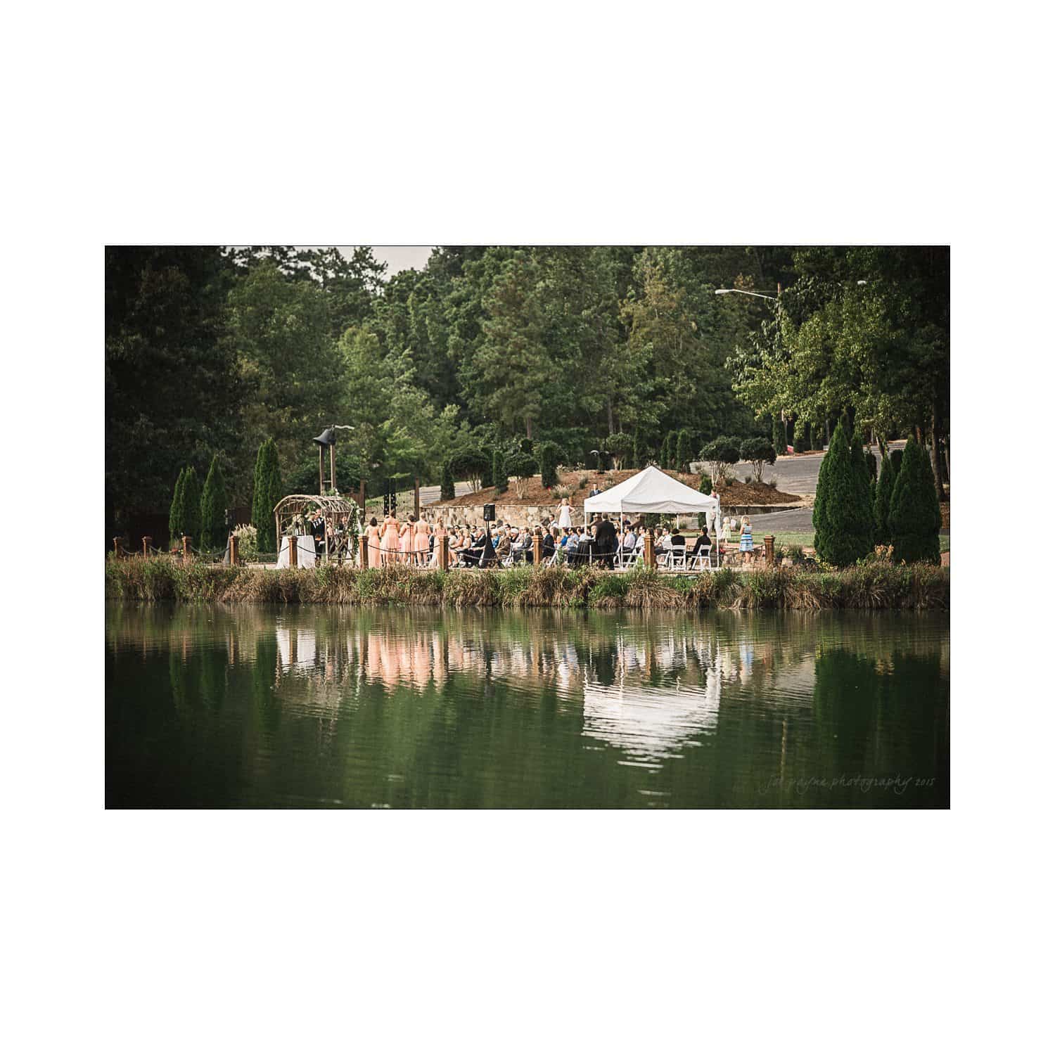 A serene lakeside wedding scene with guests seated under a white tent. The event is surrounded by lush greenery and reflected in the calm water. Outdoor decorations and seating arrangements create an elegant atmosphere.