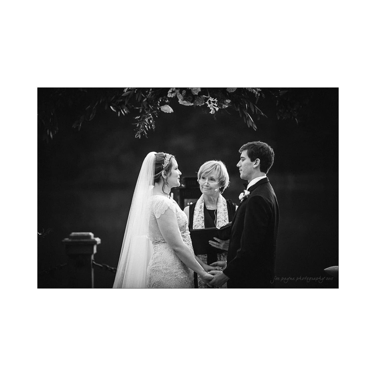A bride and groom stand facing each other, holding hands in front of an officiant during an outdoor wedding ceremony. The bride wears a veil and lace dress, while the groom is in a dark suit. A floral arch is visible in the background.