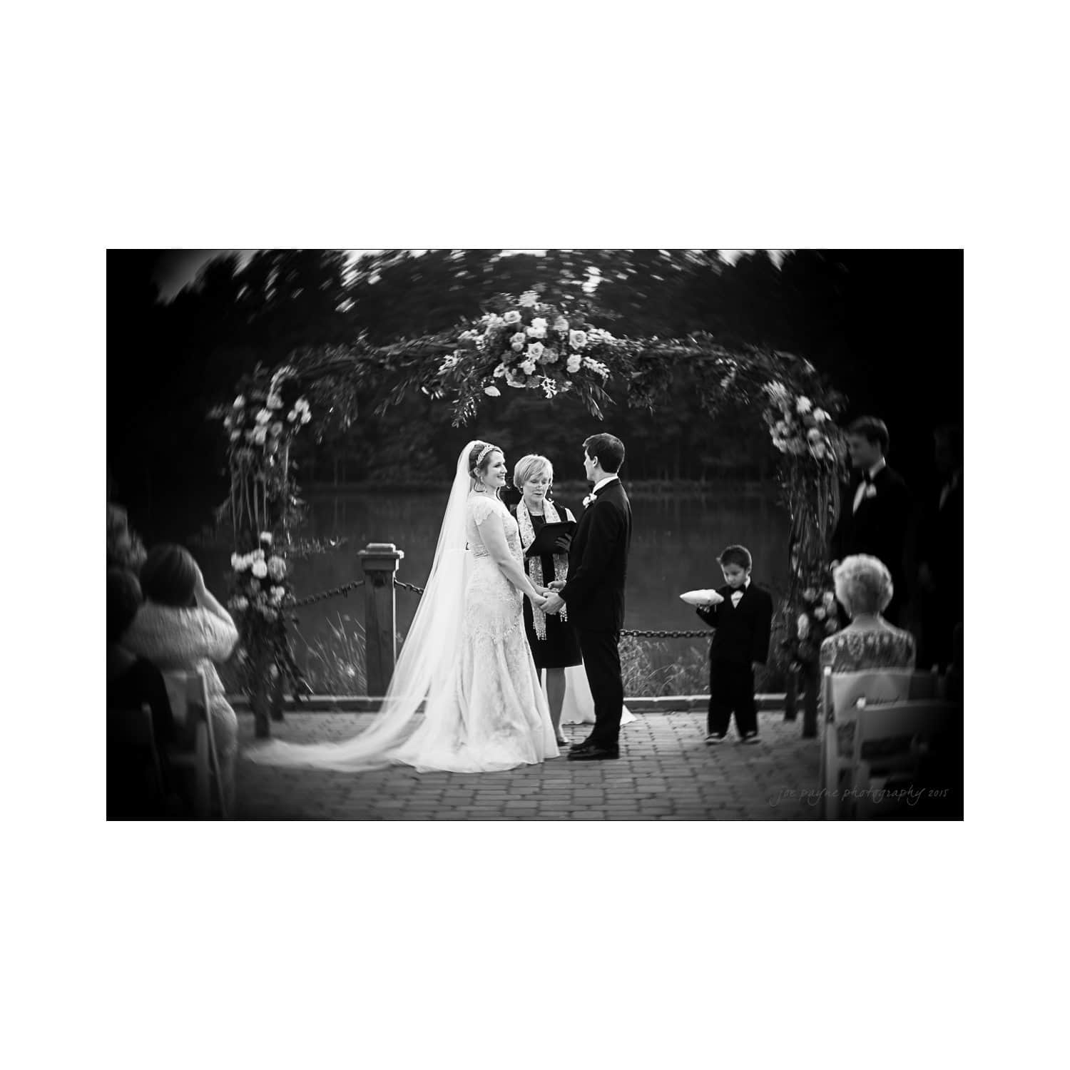 A black and white photo of a wedding ceremony by a lake. A couple stands under a floral arch, holding hands. The officiant stands behind them. Guests are seated on either side, and a child is visible to the right holding a piece of paper.