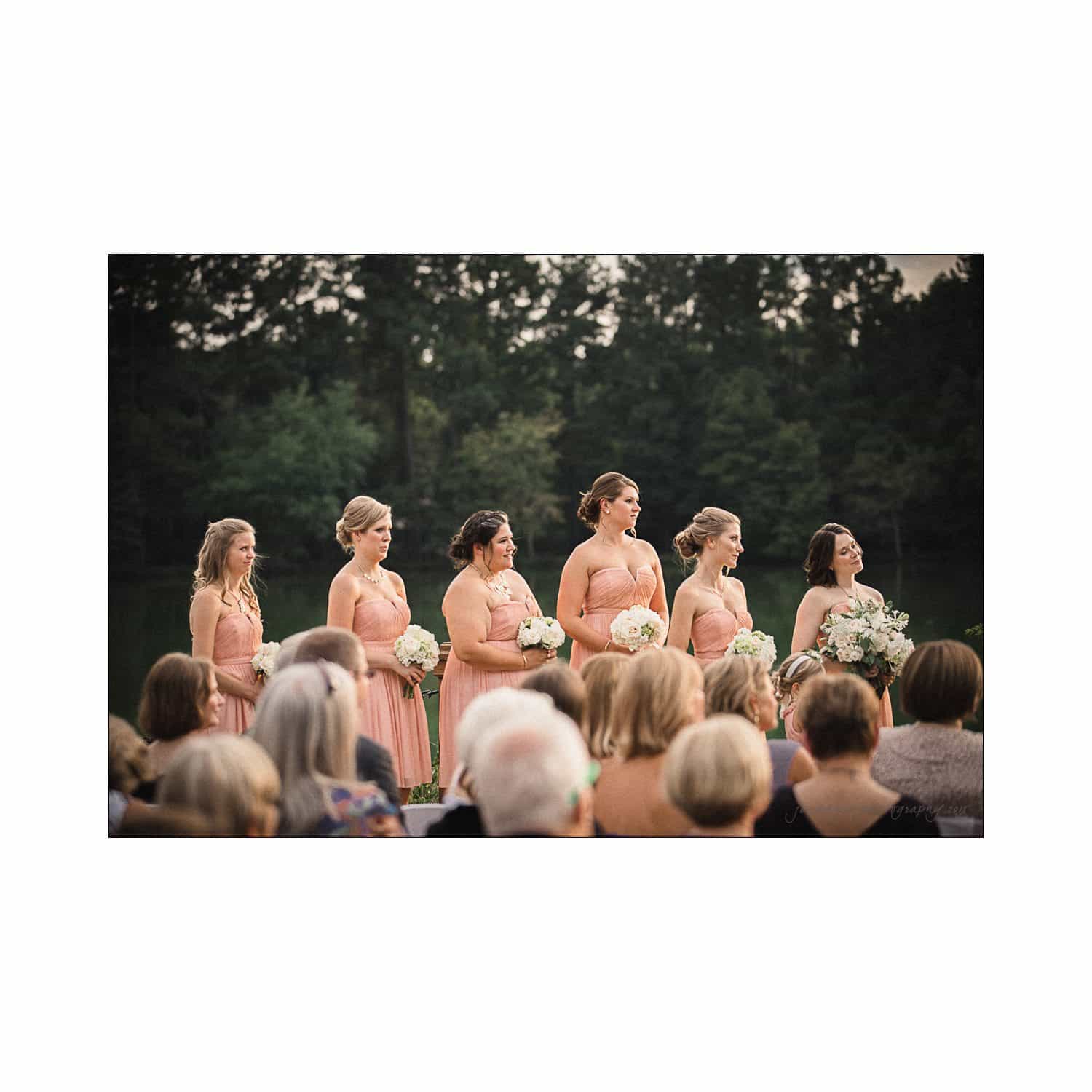 A group of bridesmaids in peach dresses stand outdoors during a wedding ceremony. They each hold bouquets and face forward. The background features lush green trees, and an audience is seated in the foreground.