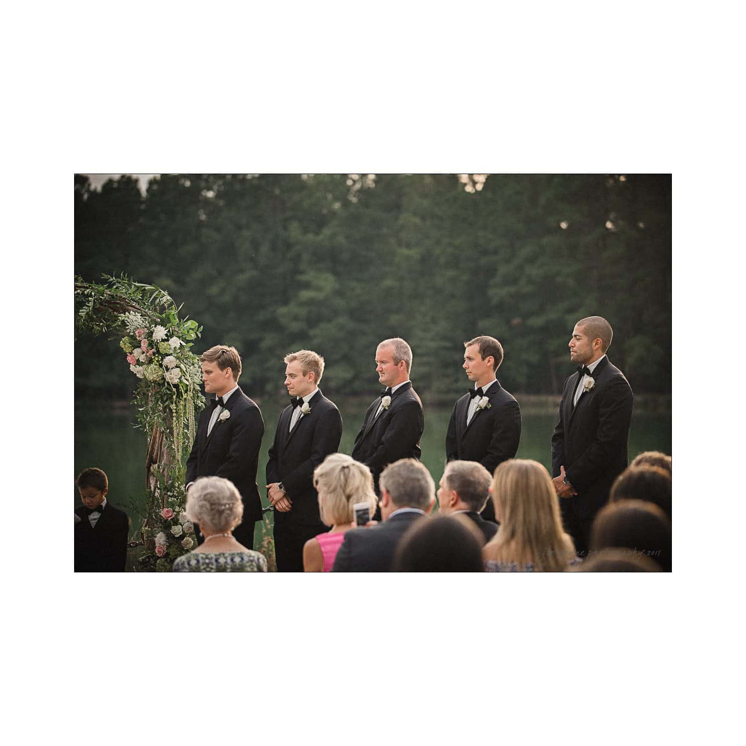 A group of groomsmen in black tuxedos stand beside a floral arch during an outdoor wedding ceremony. They look towards the ceremony, surrounded by seated guests. The background has lush green trees and a calm body of water.