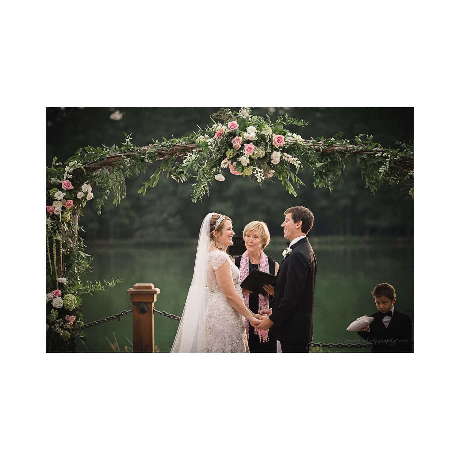 A couple in wedding attire stands under a floral arch by a lake, holding hands and smiling at each other. A celebrant stands between them, and a child peeks from behind a wooden post. The scene is serene and romantic.