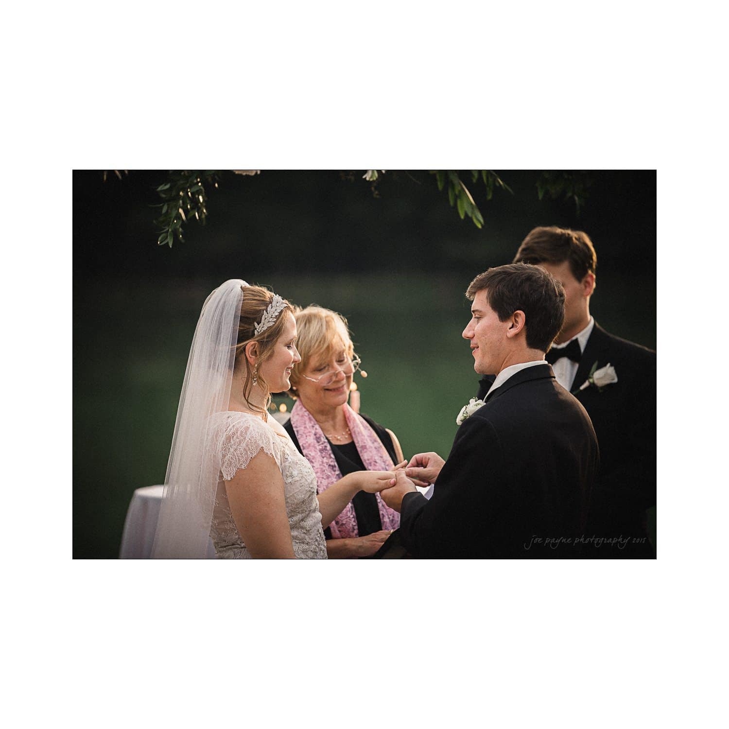 A bride and groom exchange rings during an outdoor wedding ceremony. The bride wears a white dress with a veil, and the groom is in a black suit. An officiant stands behind them, smiling. Lush greenery is visible in the background.