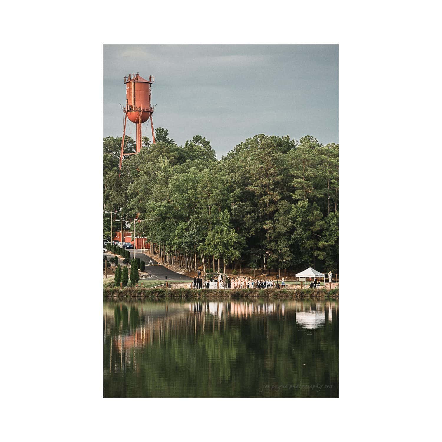 A lake reflecting a forested area with a red water tower in the background. A group of people is gathered near the shore under a small white tent. The scene is calm with a cloudy sky overhead.