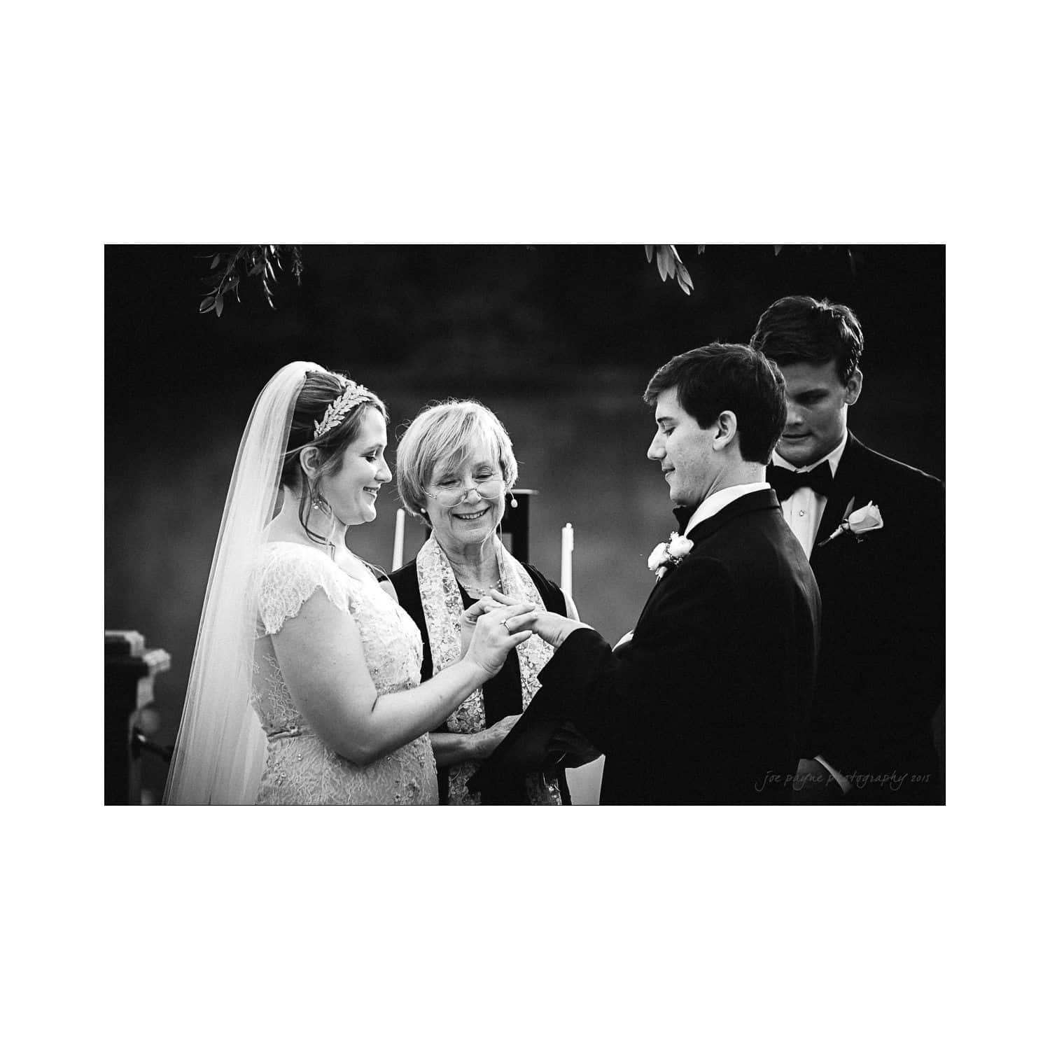A black and white photo of a wedding ceremony. A bride and groom exchange rings while an officiant stands between them. A groomsman stands beside the groom. The bride wears a veil and lace dress; the groom is in a suit.