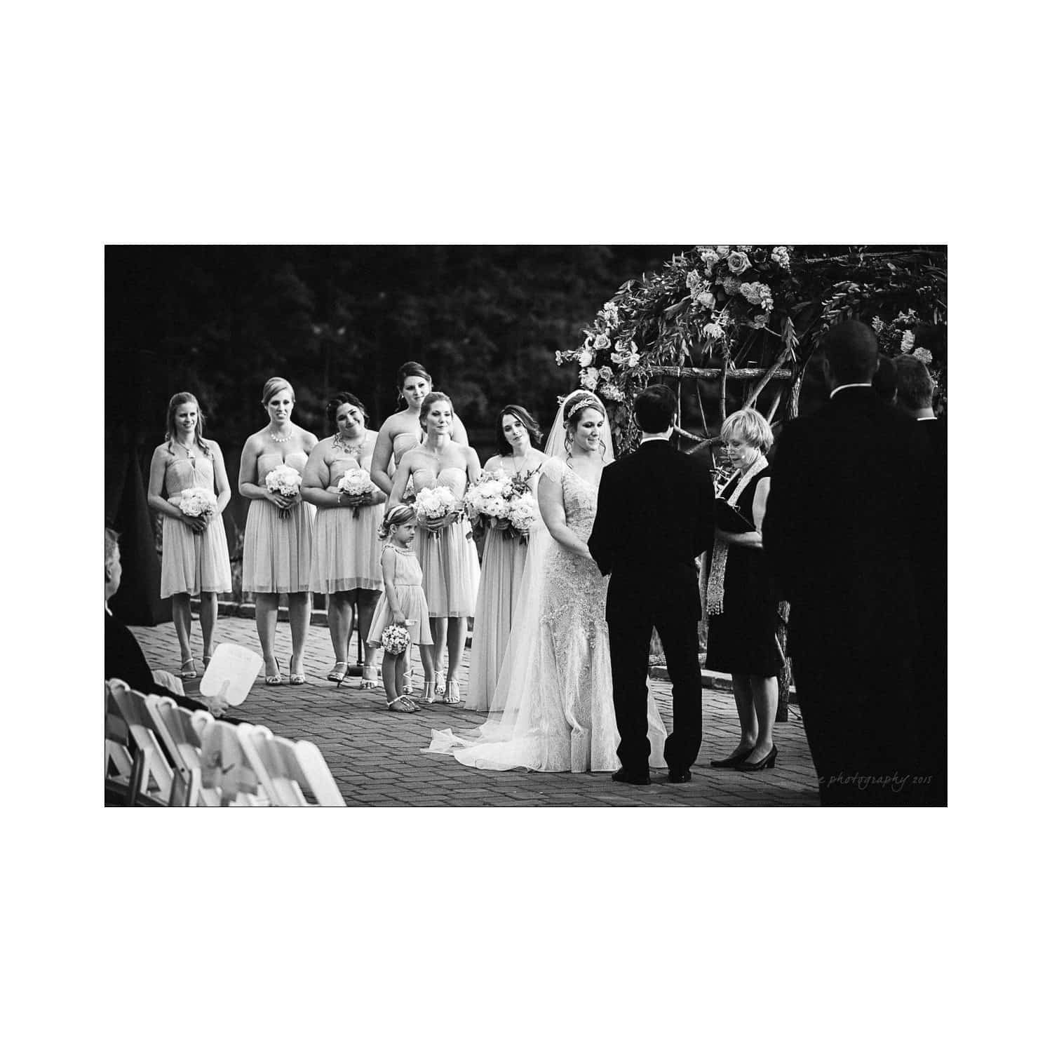 A black-and-white photo of a wedding ceremony. The bride in a long dress holds a bouquet, standing beside the groom. Bridesmaids in matching dresses stand to the side. An officiant is present, and guests are seated in rows.