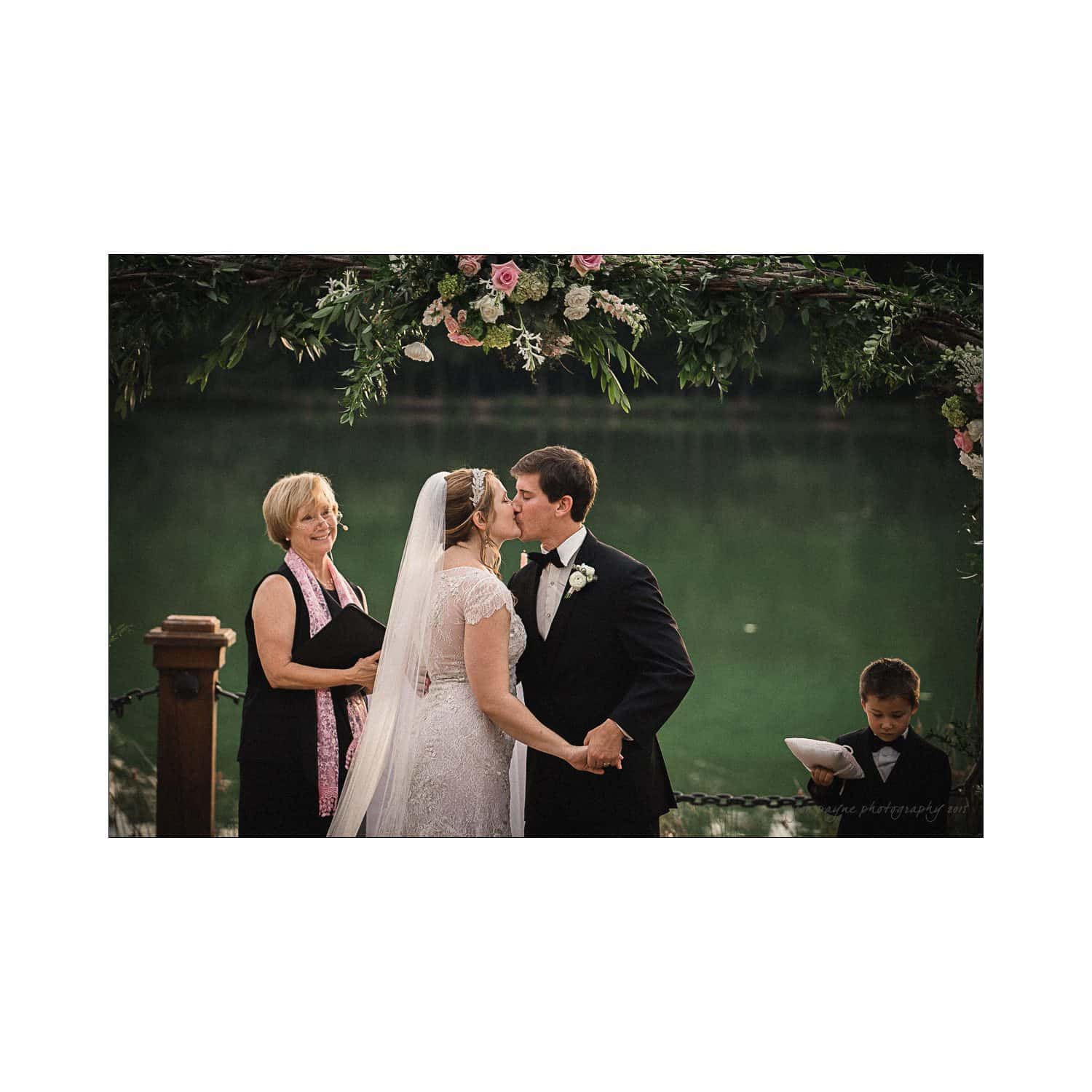 A bride and groom kiss under a floral arch by a lake during their wedding ceremony. A woman officiates on the left, and a young ring bearer stands on the right holding a pillow. Lush greenery surrounds the serene setting.