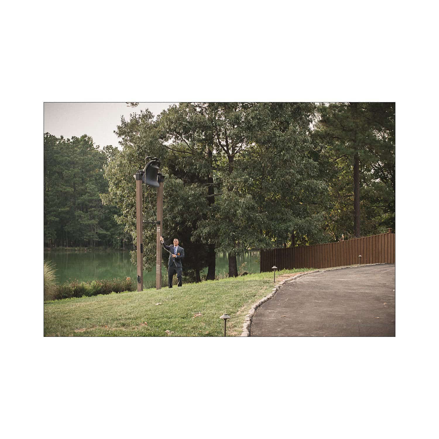 A person ringing a large bell mounted on a wooden post near a walkway. The area is grassy and surrounded by trees, with a lake visible in the background. The scene conveys a peaceful, outdoor setting.