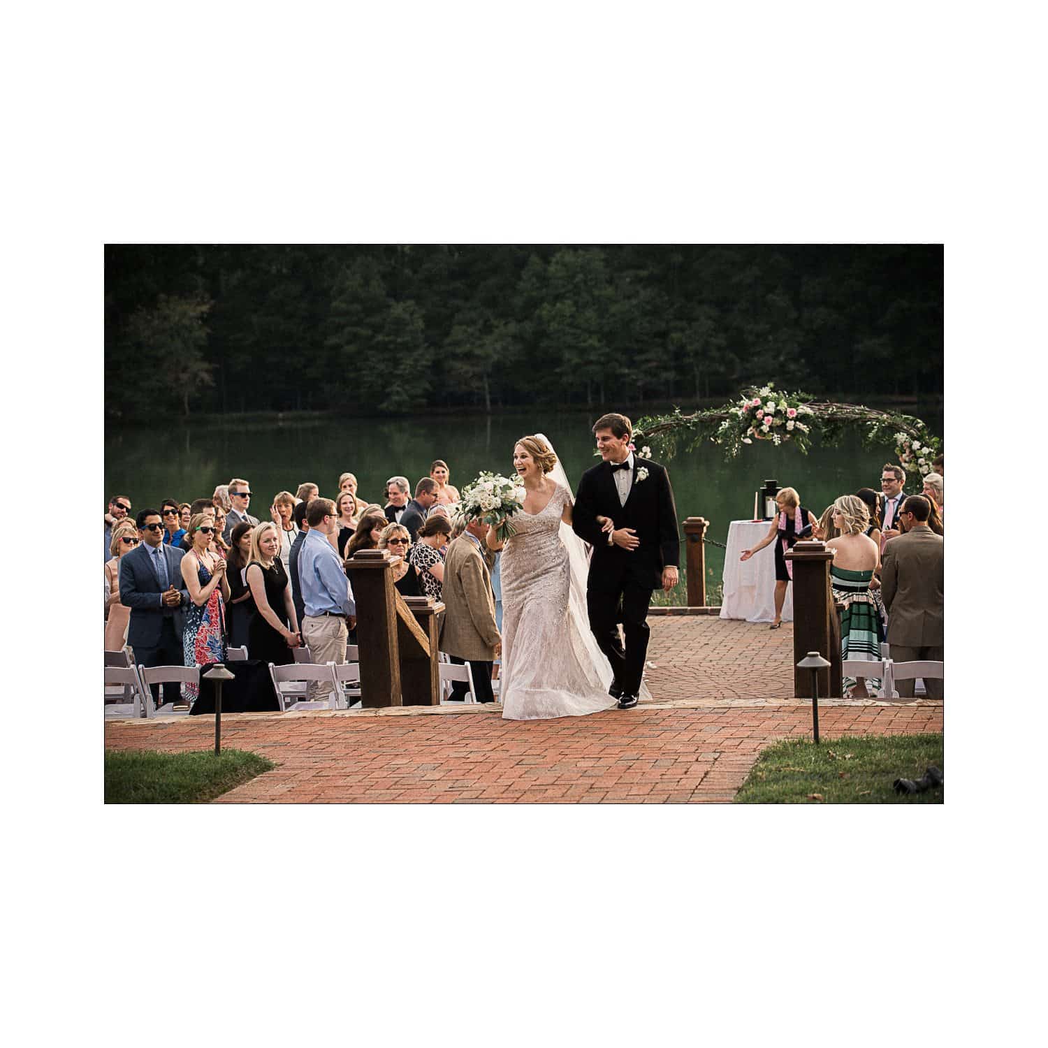 A newlywed couple walks down an outdoor aisle after their ceremony. The bride is in a white dress holding a bouquet, and the groom is in a black suit. Guests are seated on either side, and a scenic lake is visible in the background.
