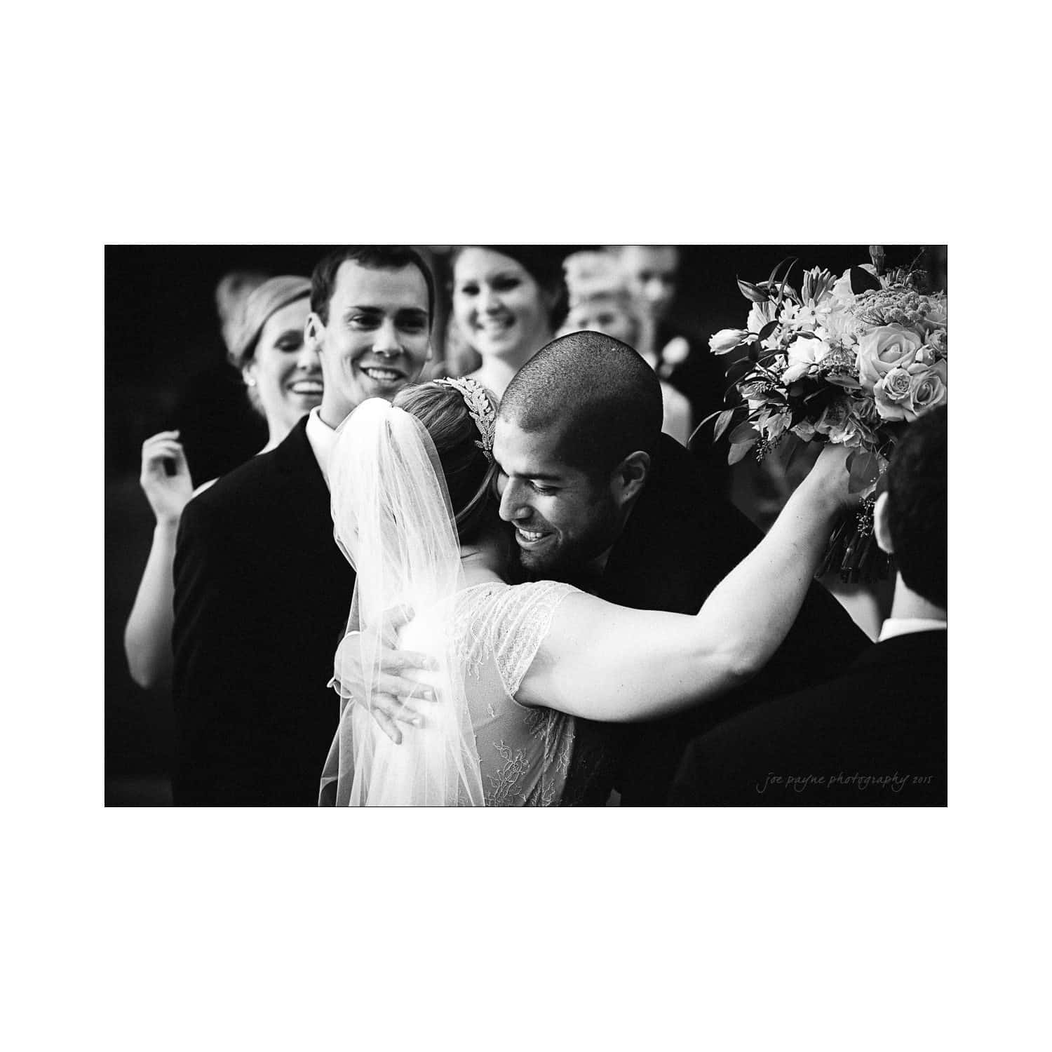 A bride and groom embrace joyfully amidst a celebrating wedding party. The bride holds a bouquet of flowers, while several people in the background smile and share in the moment. The image is in black and white.
