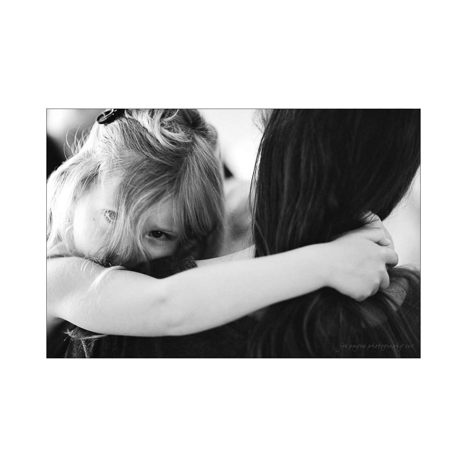 A young child with light hair peeks over a womans shoulder, embracing her in a close hug. The image is in black and white, capturing a moment of tenderness and connection.