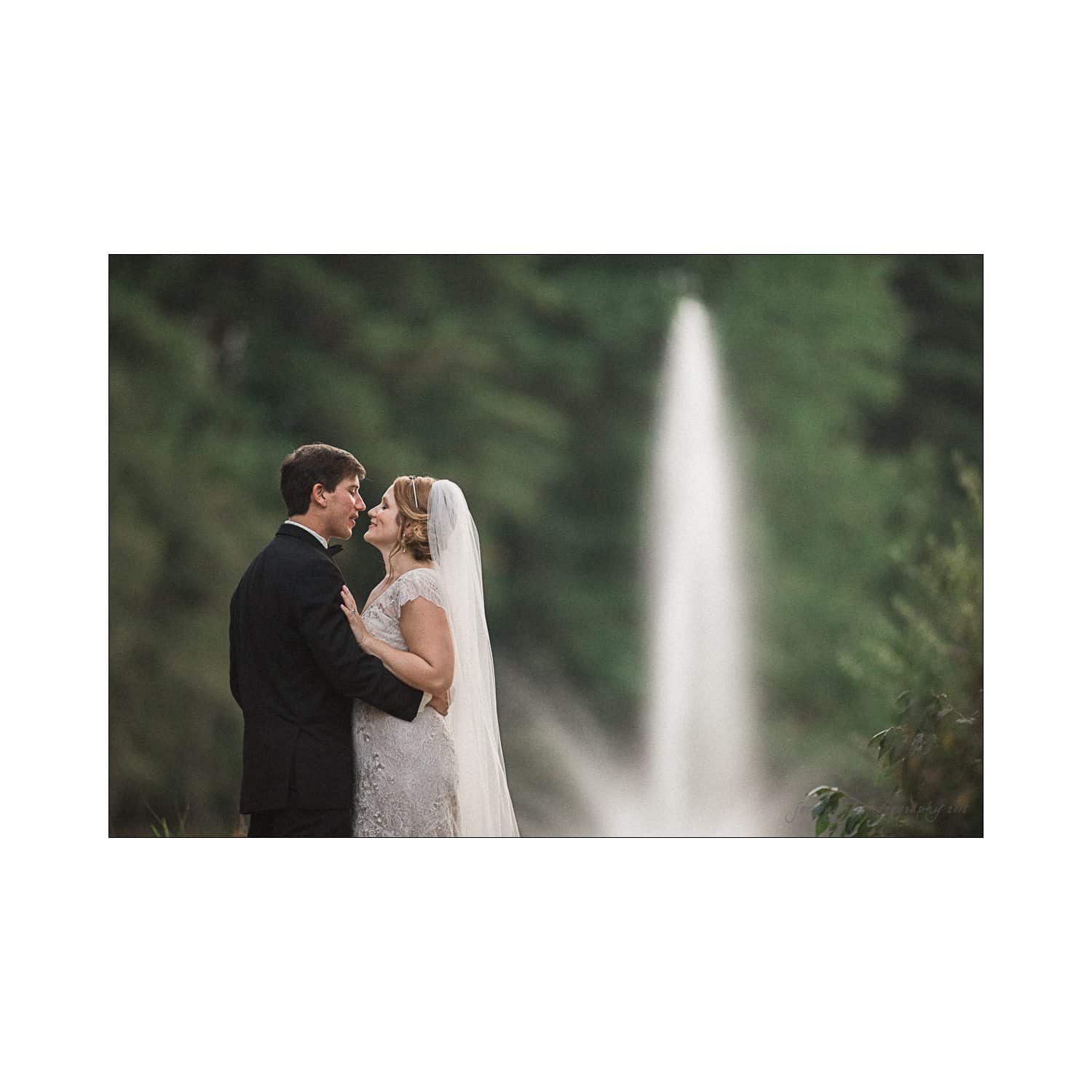 A bride and groom gaze at each other while embracing, standing in front of a tall fountain. The bride wears a lace gown with a veil, and the groom is in a dark suit. Lush greenery surrounds the scene, creating a serene, romantic atmosphere.