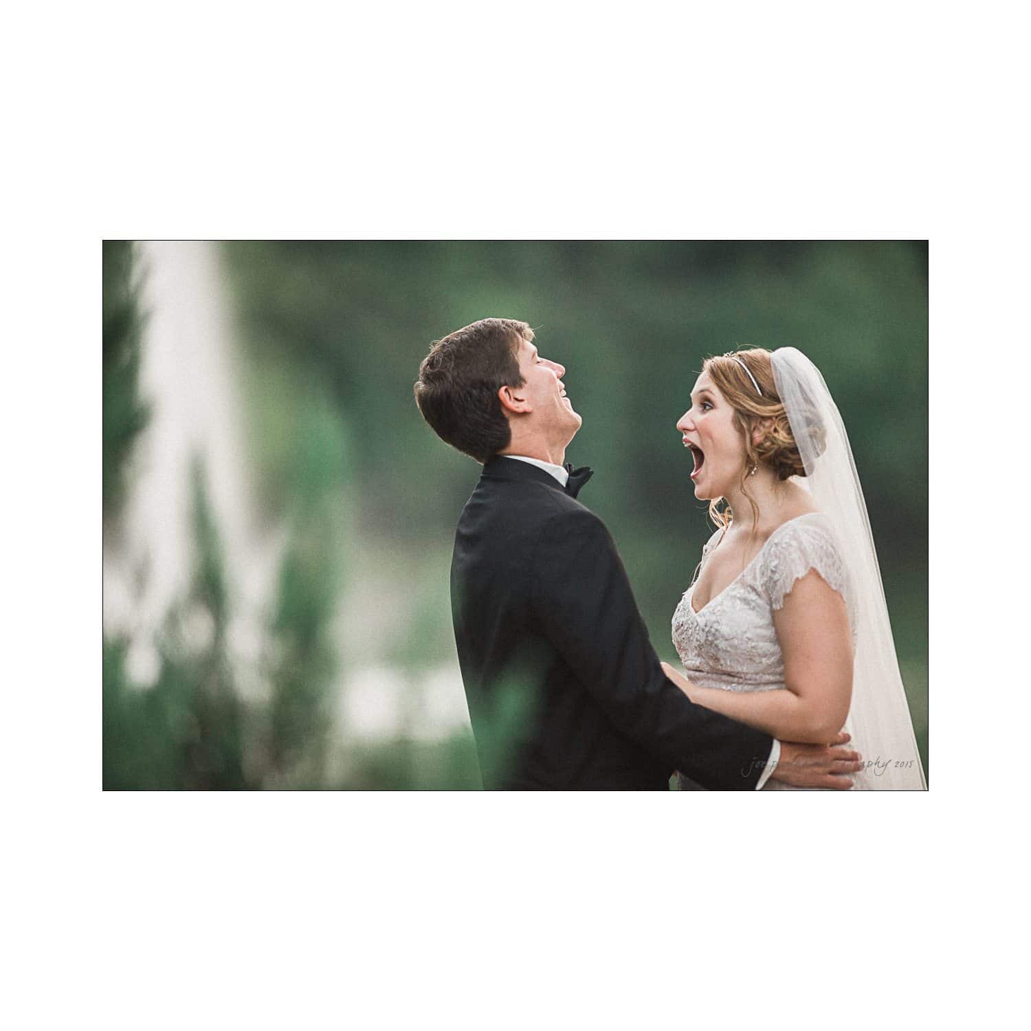 Bride and groom share a joyful and surprised moment outdoors. The bride wears a lace gown and veil, while the groom is in a black tuxedo. They are laughing together with greenery in the background.