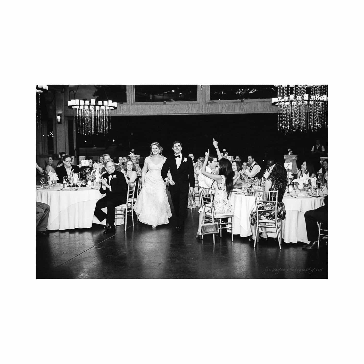 A bride and groom walk hand in hand into a wedding reception. Guests sit at round tables, some clapping and cheering. The venue is elegantly decorated with chandeliers. The image is in black and white.