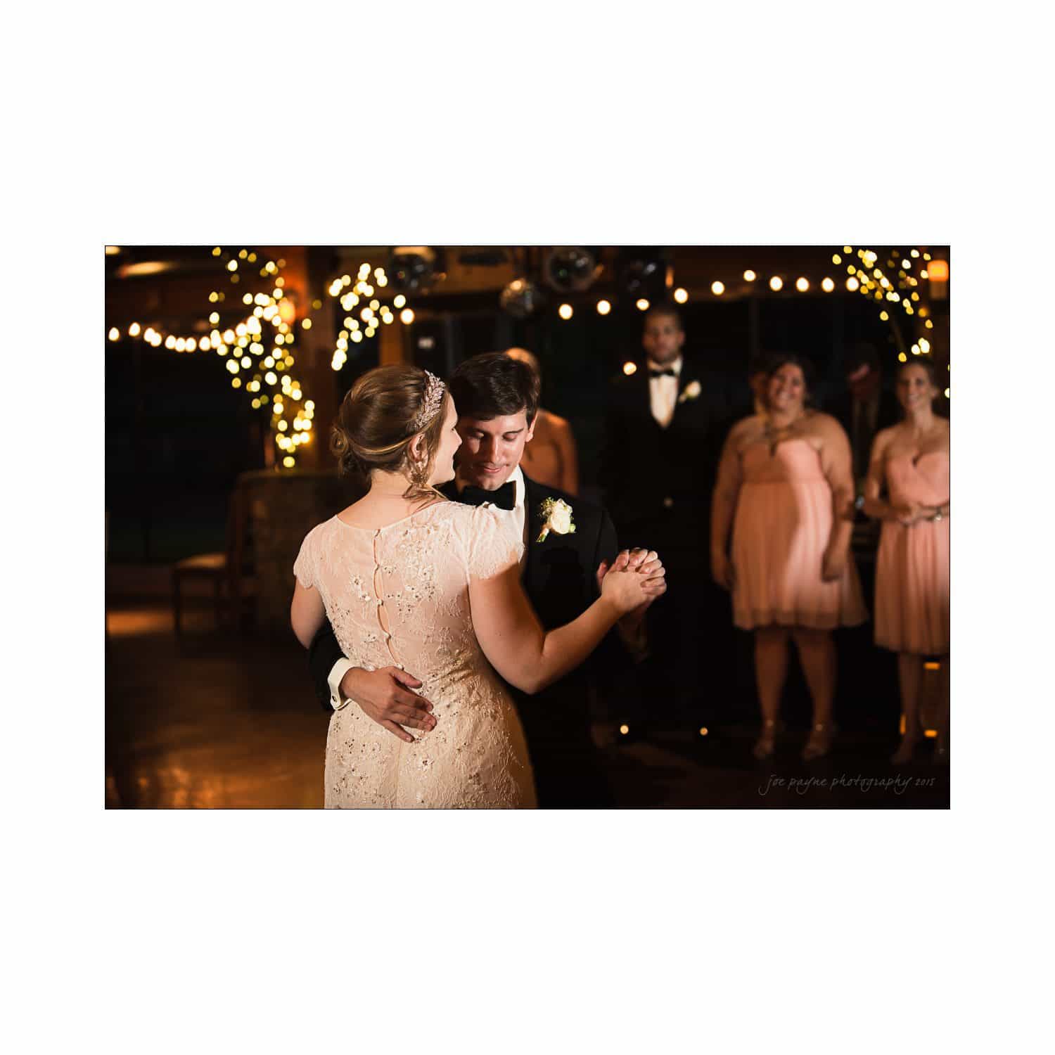 A couple shares a close dance at a wedding reception. The woman wears a lace dress and the man is in a suit. Soft lighting and string lights in the background create a romantic ambiance, with the wedding party smiling in the background.