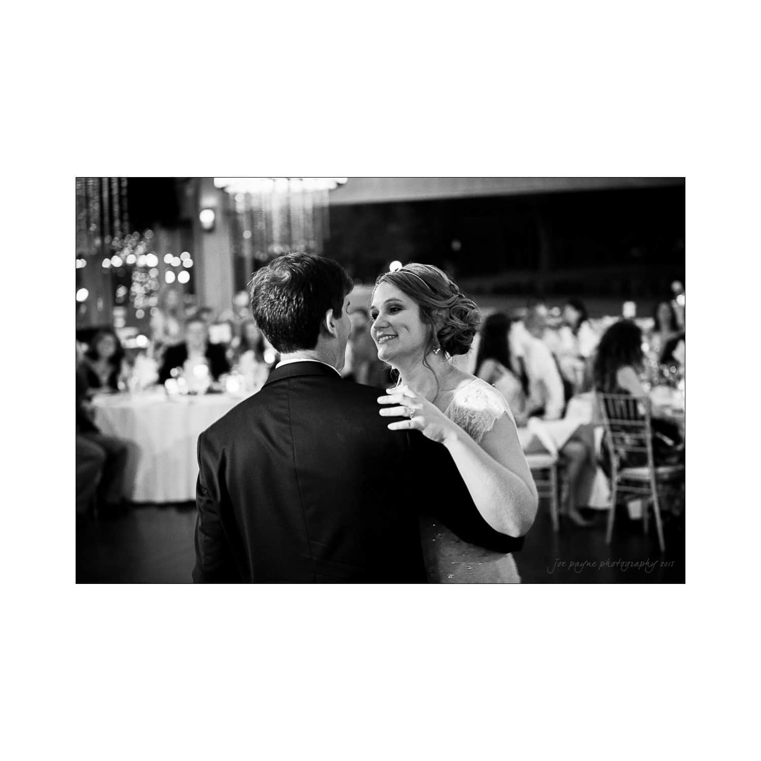 A black and white photo of a couple dancing at a wedding reception. The bride is smiling, wearing a lace dress and tiara, while the groom is in a dark suit. Guests are seated at tables adorned with white tablecloths and string lights.
