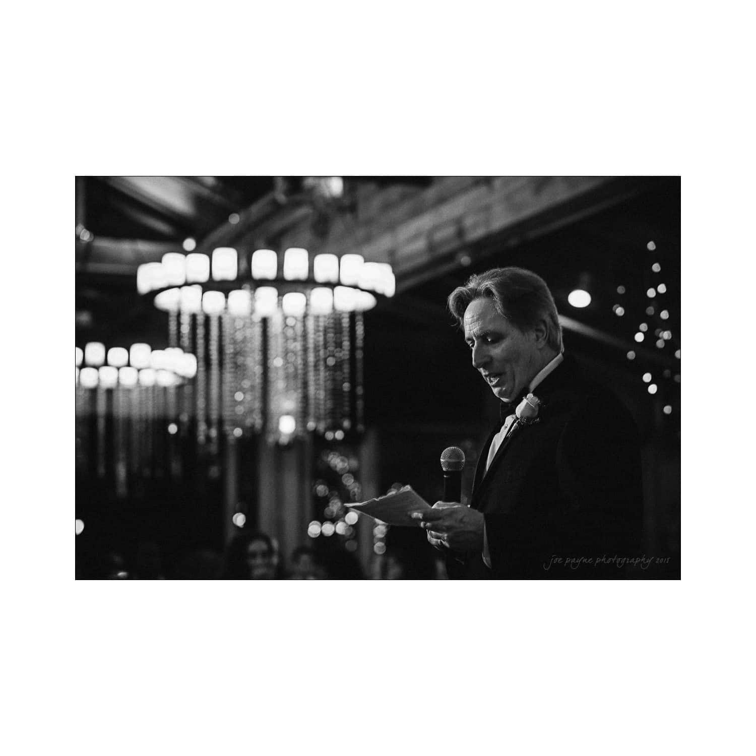 A black-and-white photo of a man in a suit speaking into a microphone at an indoor event. He holds a folded piece of paper. Chandelier lights and blurred guests are visible in the background.