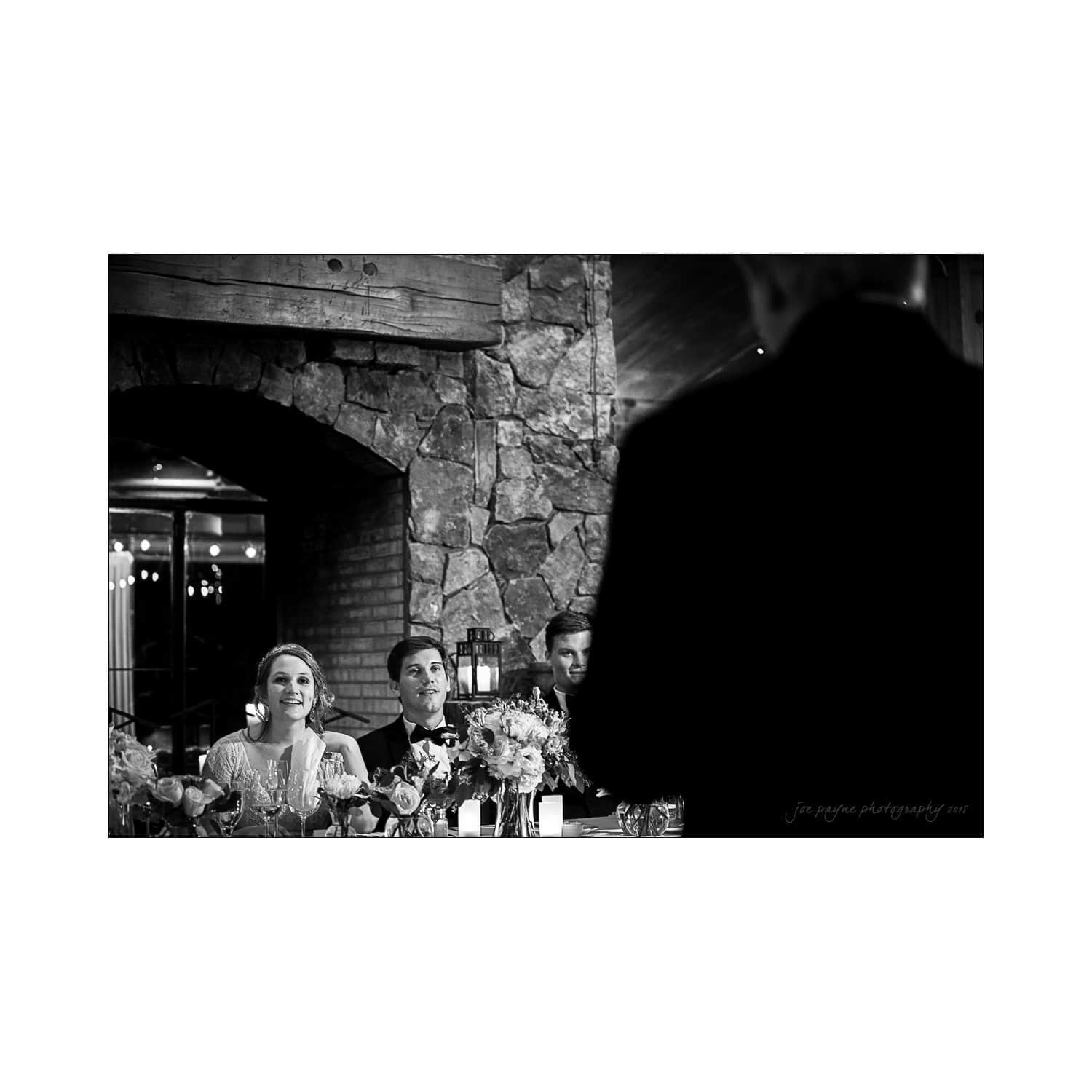 A black and white photo of a wedding reception. A bride and groom sit at a table smiling, looking towards a speaker who is standing in the foreground. The setting is a rustic venue with stone walls and floral decorations.
