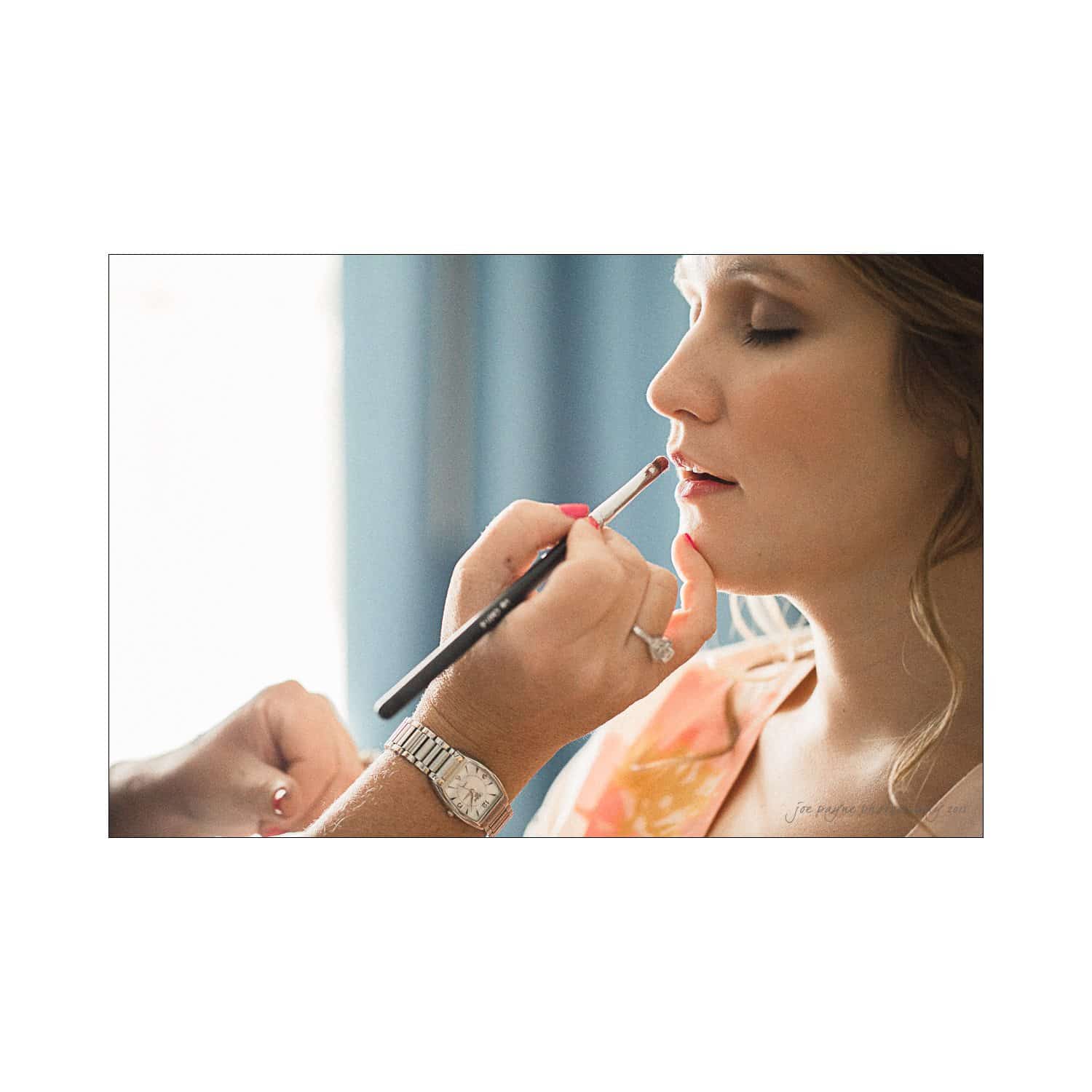 A person is having makeup applied by a makeup artist using a brush. The person is seated in profile, and the setting appears calm and focused on the makeup process.