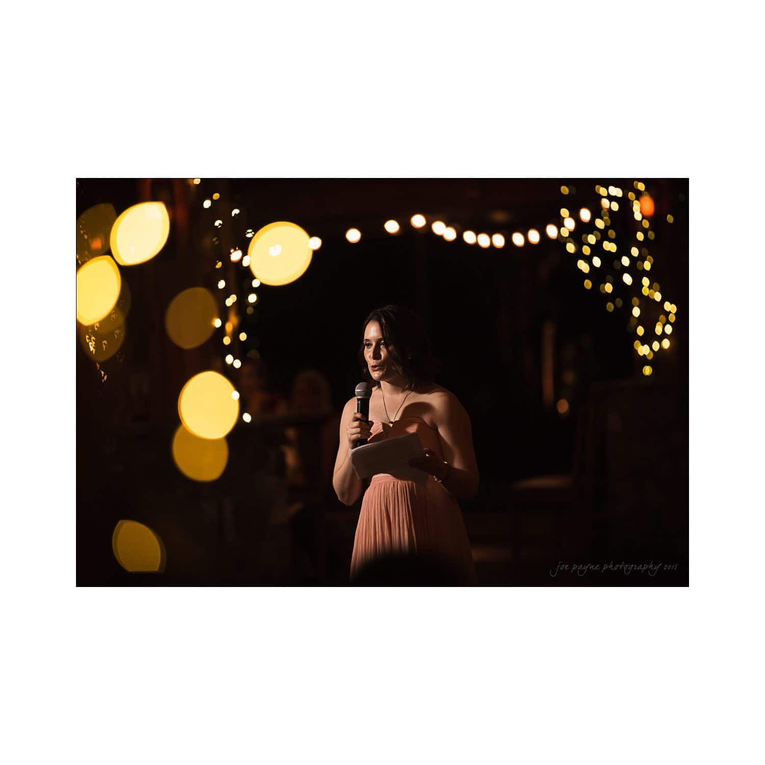 A woman in a peach dress is holding a microphone and speaking at an event. She stands in a warmly lit space adorned with round string lights.