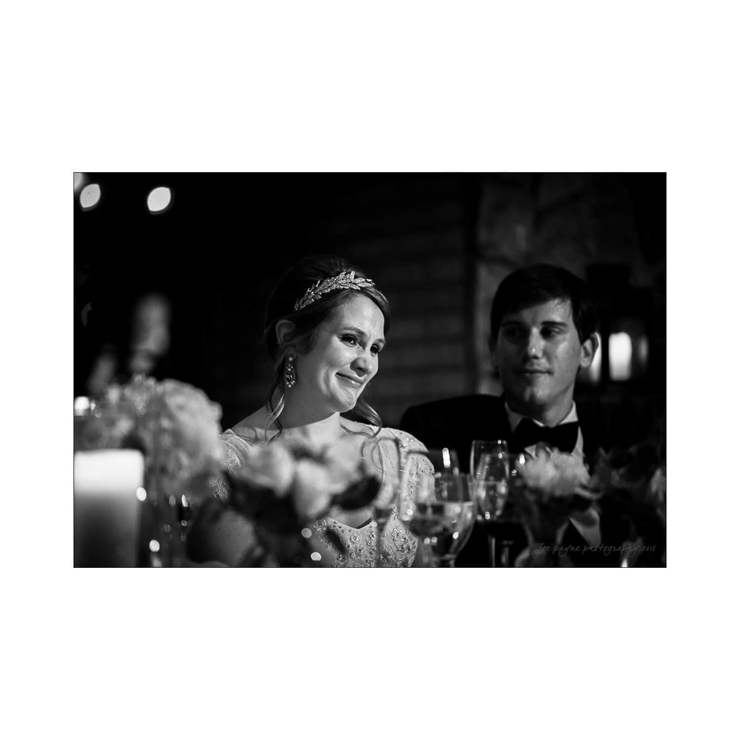 A black-and-white photo of a wedding reception. A bride in a tiara and earrings smiles as she sits next to a man in formal attire. They are surrounded by floral arrangements and lit candles, creating an intimate atmosphere.