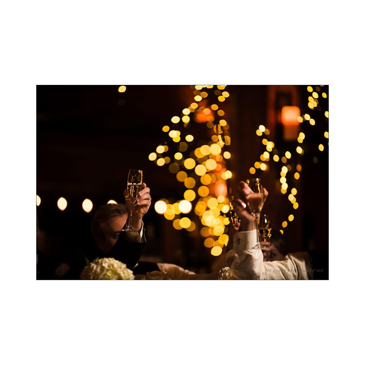Hands raise glasses in a toast at a celebratory event. The background is softly blurred with warm, glowing bokeh lights, creating a festive and joyful atmosphere.