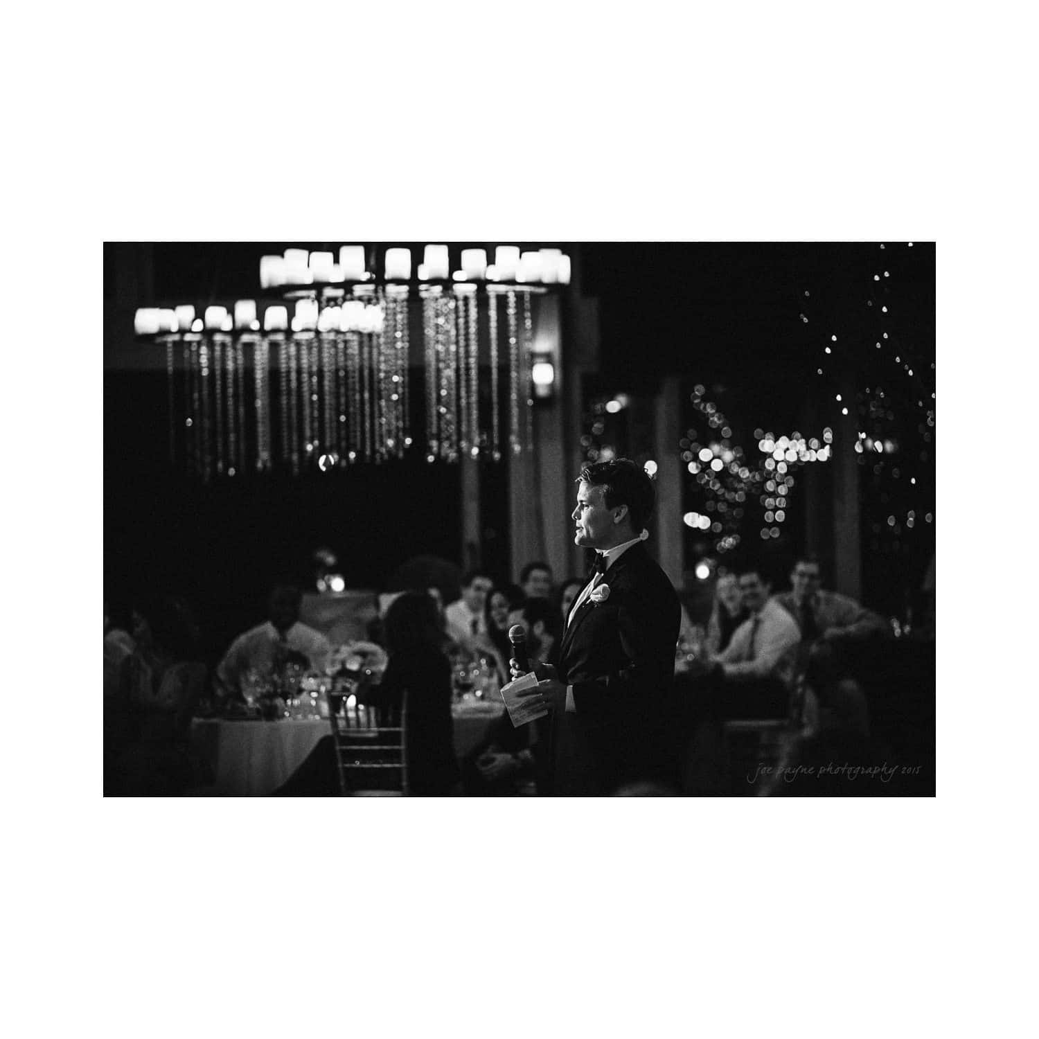 A black and white photo of a man in a tuxedo speaking at a formal event. He stands under a large chandelier, and guests sit at tables decorated with lights in the background, creating an elegant atmosphere.