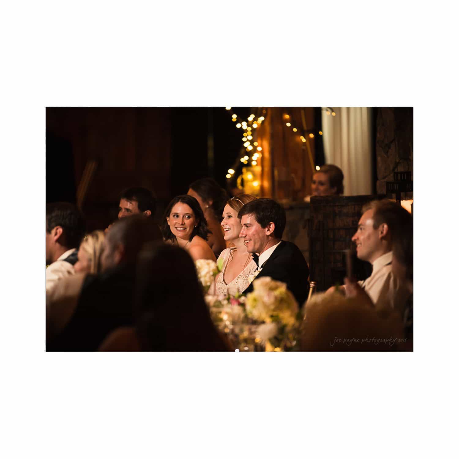 A group of people dressed formally sits at a table, smiling and engaged at what seems to be a wedding celebration. Soft, warm lighting and decorative string lights create a festive, intimate atmosphere.