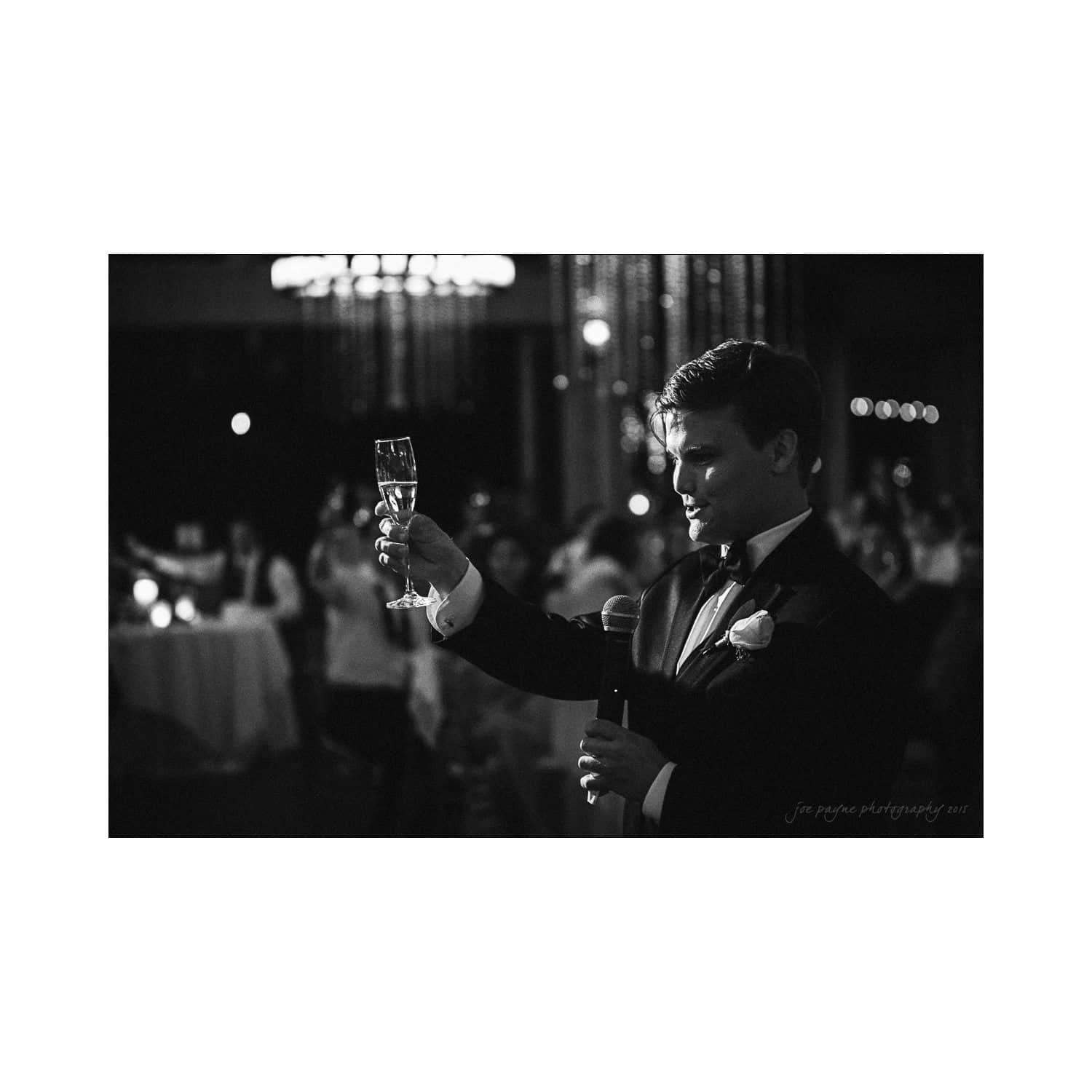 A person in a formal suit holds a microphone and raises a champagne glass in a toast during an event. The background is softly blurred, suggesting a festive atmosphere with several people seated at tables.