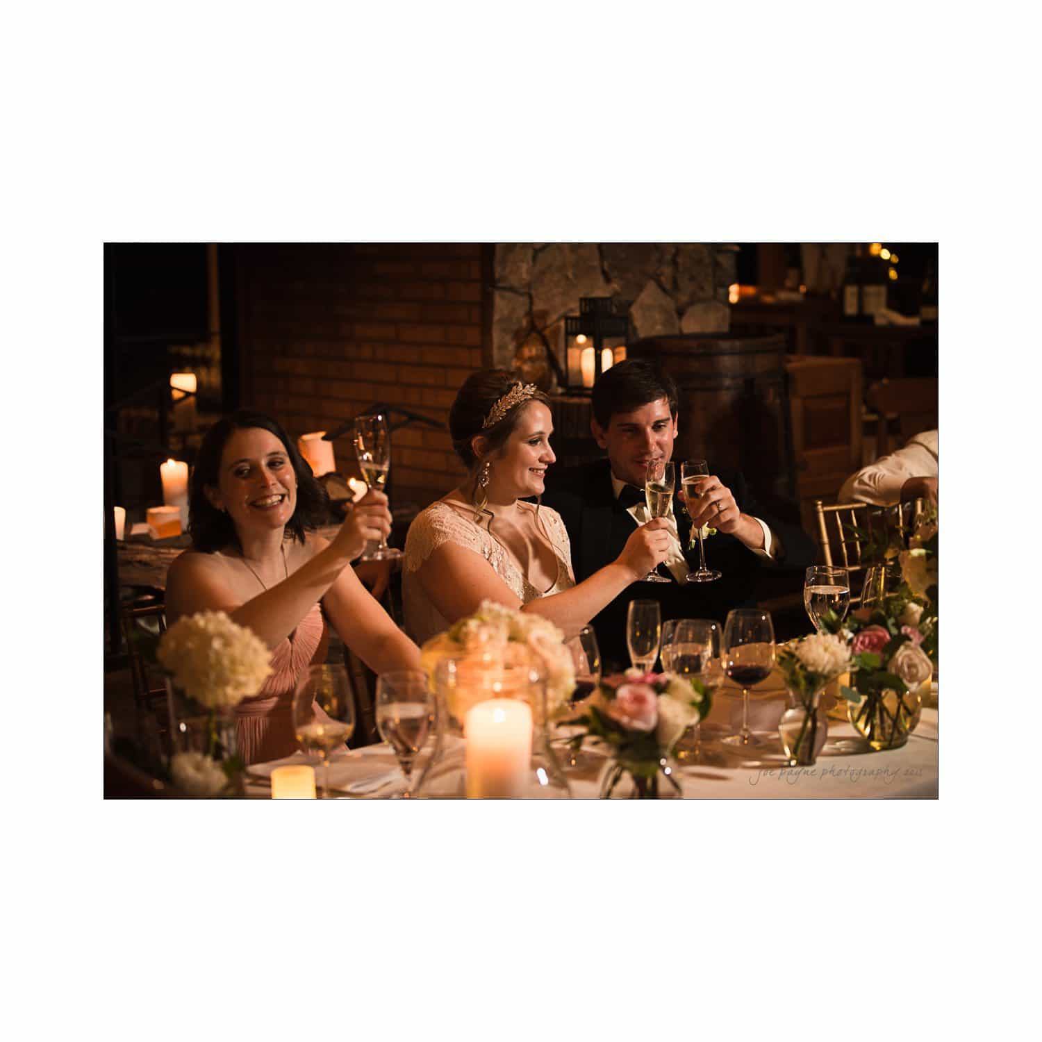 Three people dressed formally sit at a dinner table, smiling and raising glasses in a toast. The table is elegantly set with candles and floral arrangements, creating a warm and festive ambiance.