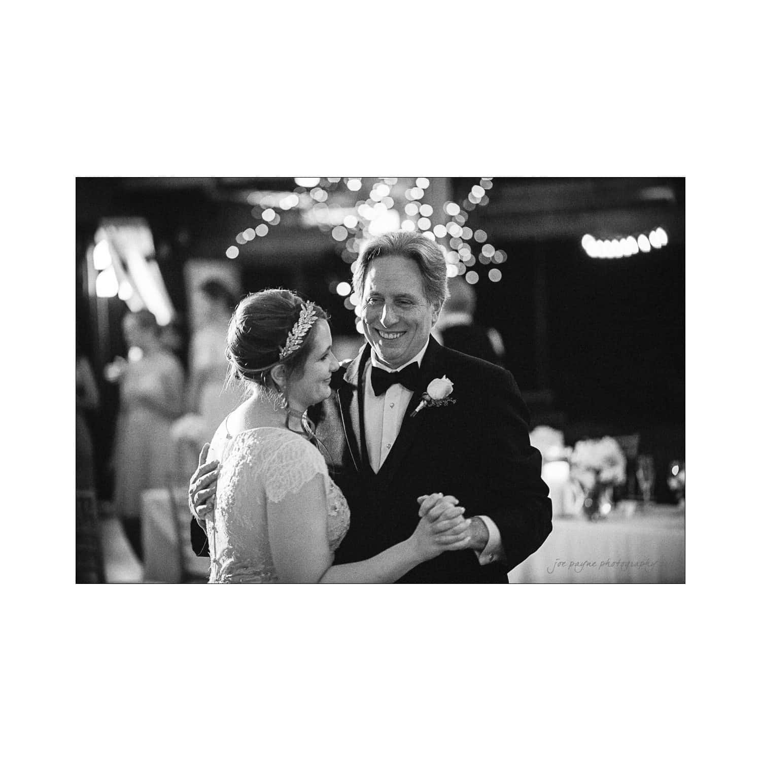 A black and white photo of a couple dancing at an event. The woman is wearing a lace dress with a headpiece, and the man is in a tuxedo with a boutonniere. They are smiling joyfully, with blurred lights and people in the background.