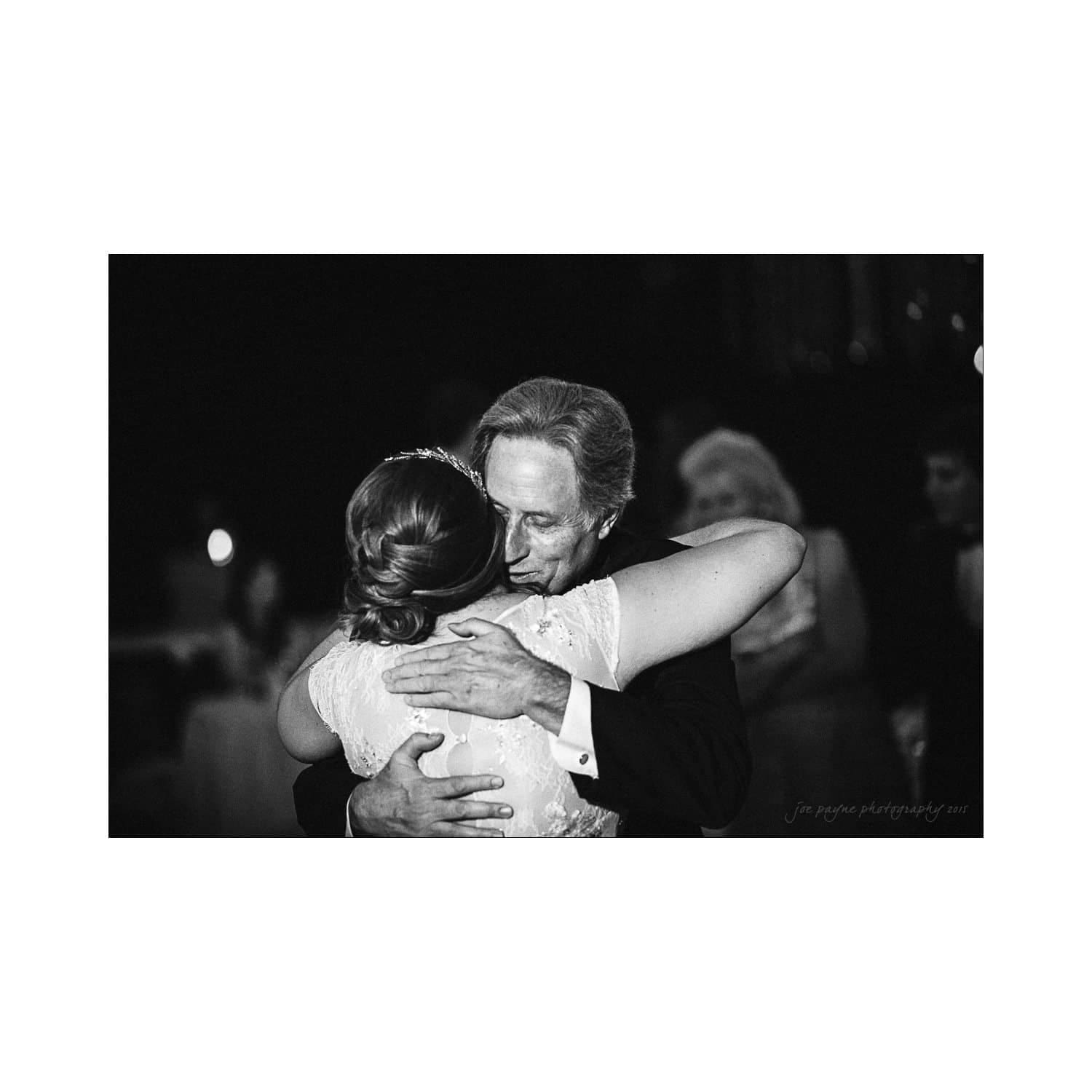 A black-and-white image of a couple embracing closely in a dance. The man, in a dark suit, is holding the woman, who is wearing a light-colored dress. The background is softly blurred, focusing on their intimate moment.