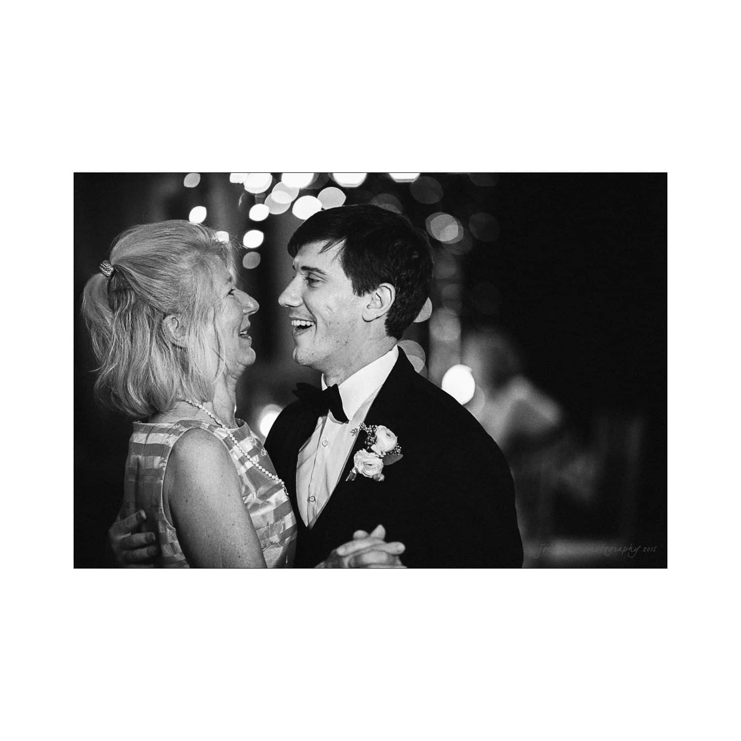A black and white photo of a man in a suit and bow tie dancing with an older woman in a striped dress. They are smiling at each other, surrounded by blurred lights in the background.