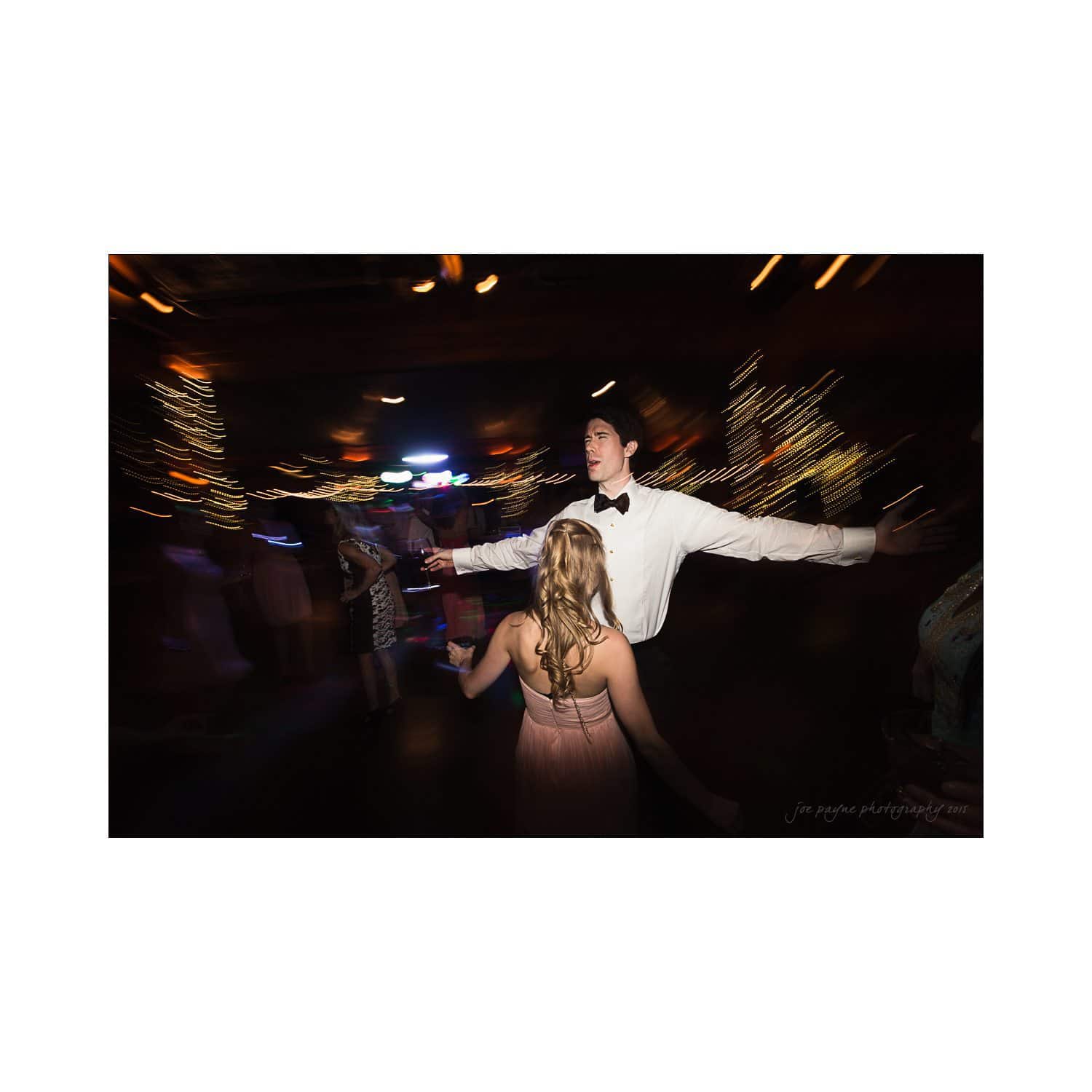 A man in a white shirt and bow tie stands with arms outstretched, facing a woman with blonde hair in a formal dress. The background is blurred, featuring colorful lights and other people dancing.