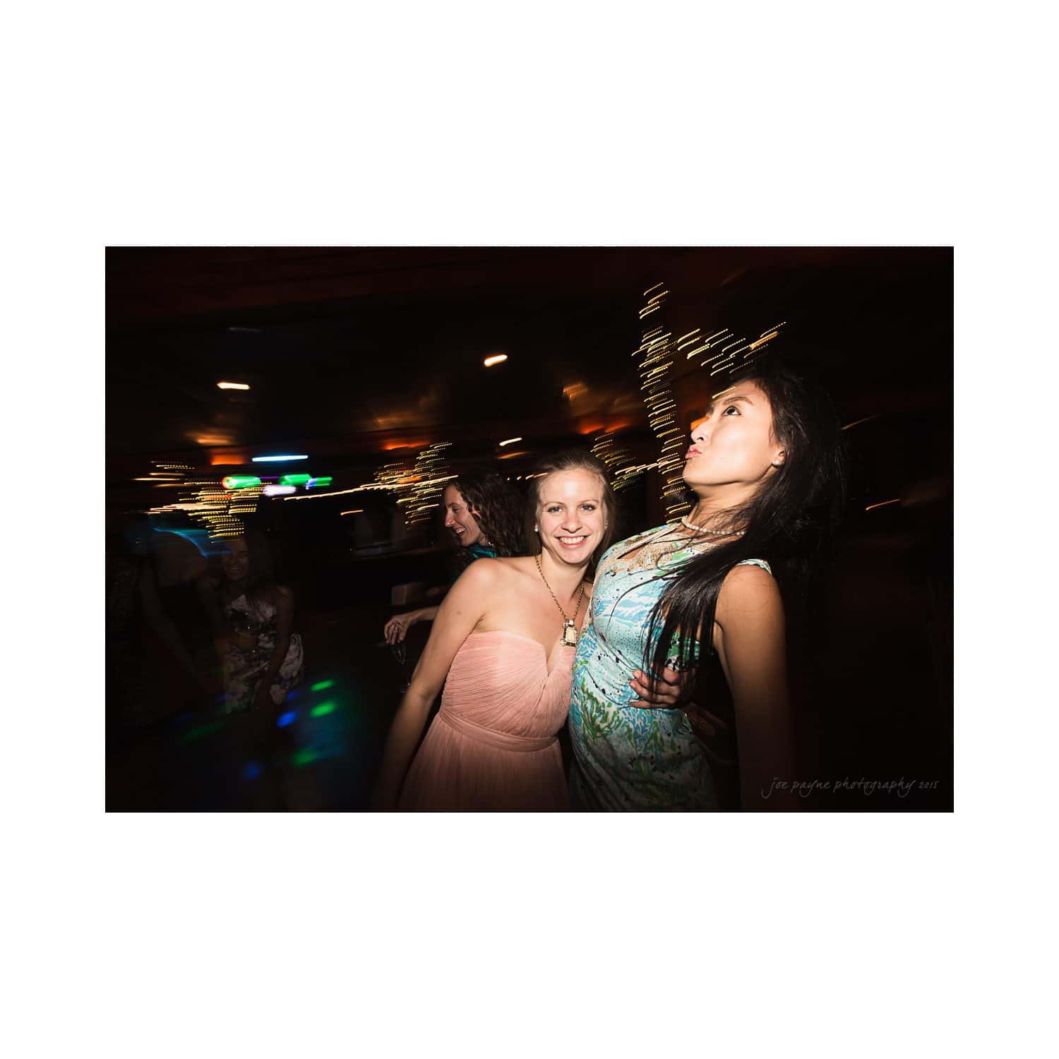 A group of women are enjoying a night out, dancing together with smiles. The background is blurred, with twinkling lights and a lively atmosphere.