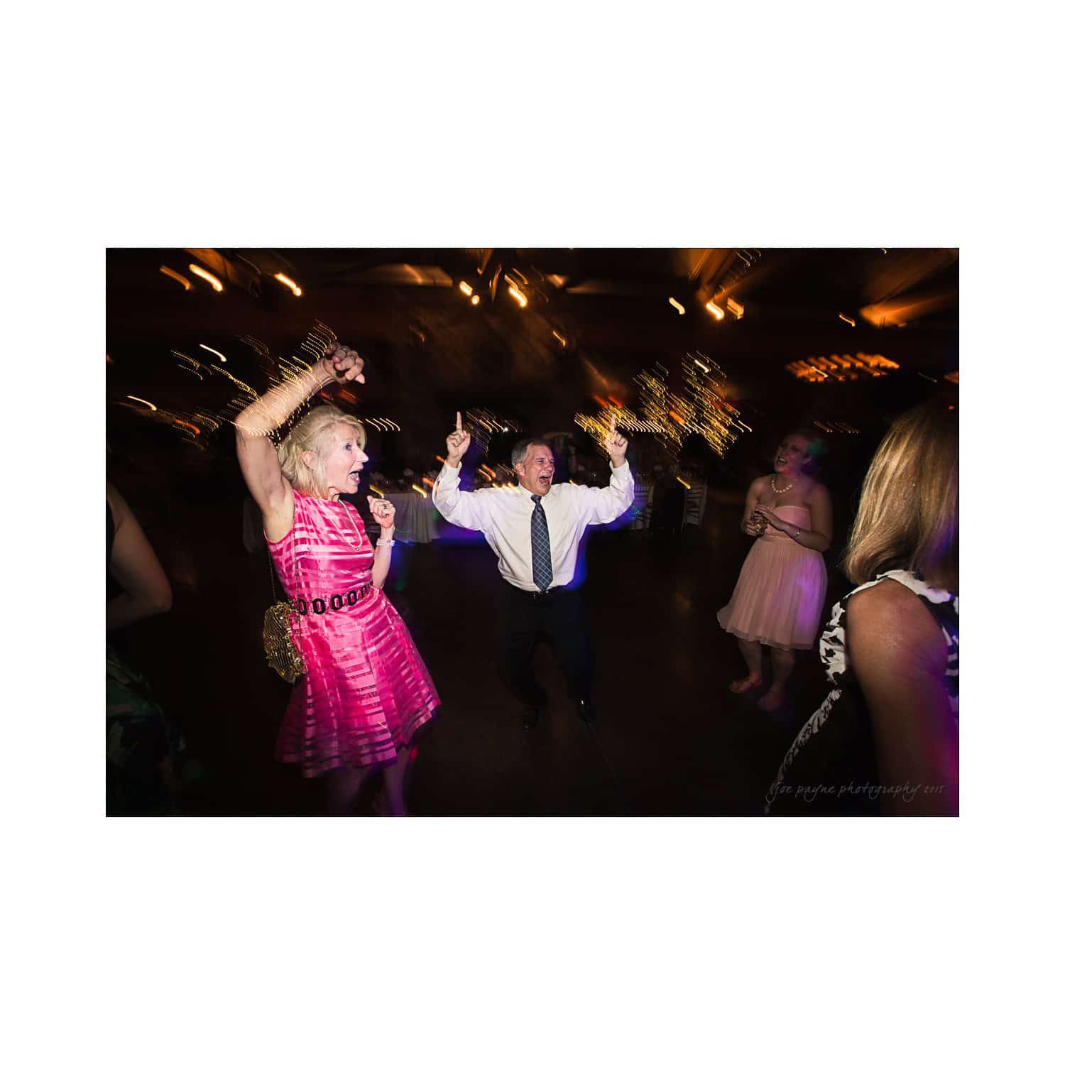 People dancing energetically at a party with festive lighting. A woman in a pink dress raises her arms, while a man in a tie dances exuberantly in the center, surrounded by other partygoers. The scene is vibrant and lively.