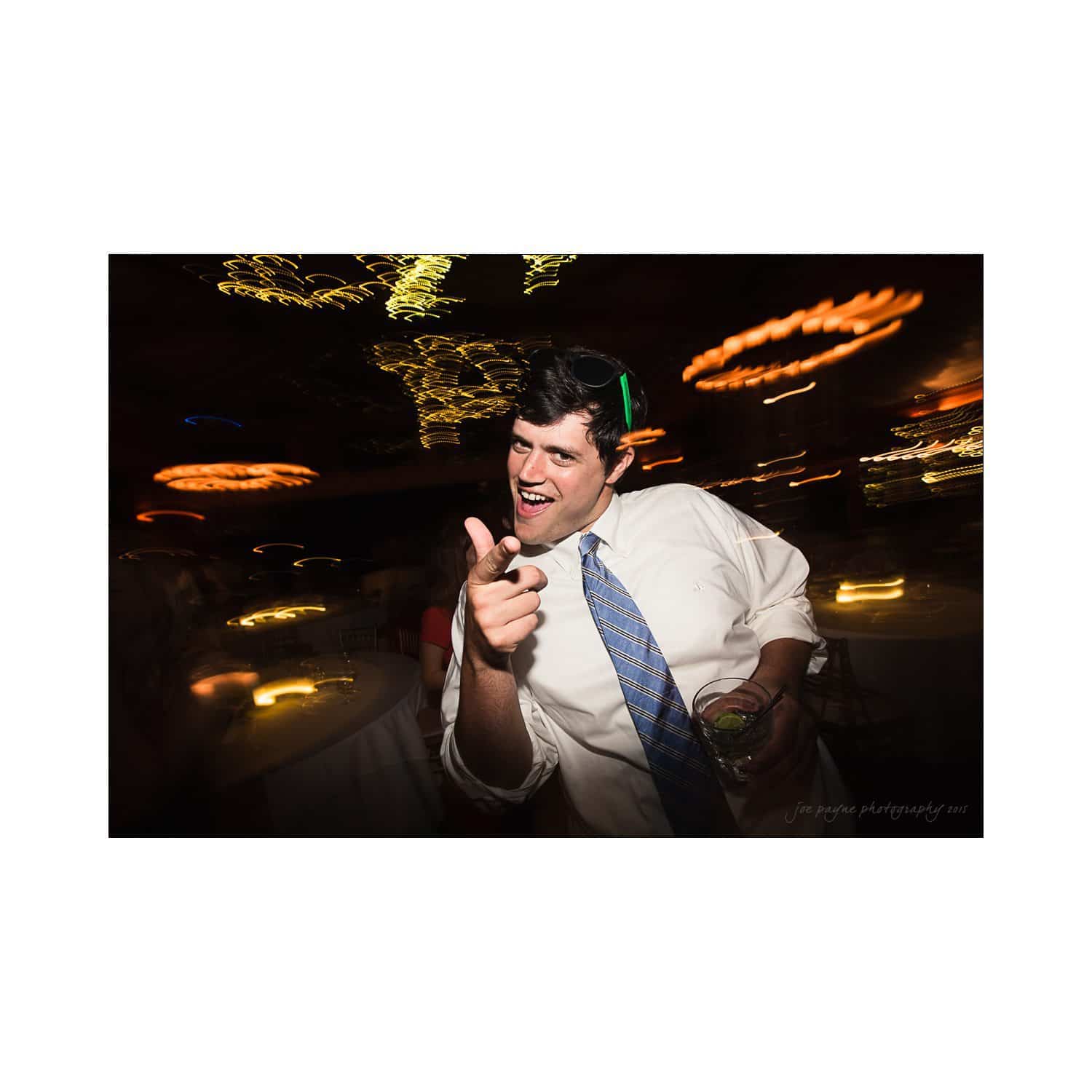 A man in a white shirt and blue tie is pointing at the camera with a playful expression. The background is blurred with colorful lights, suggesting a lively setting.