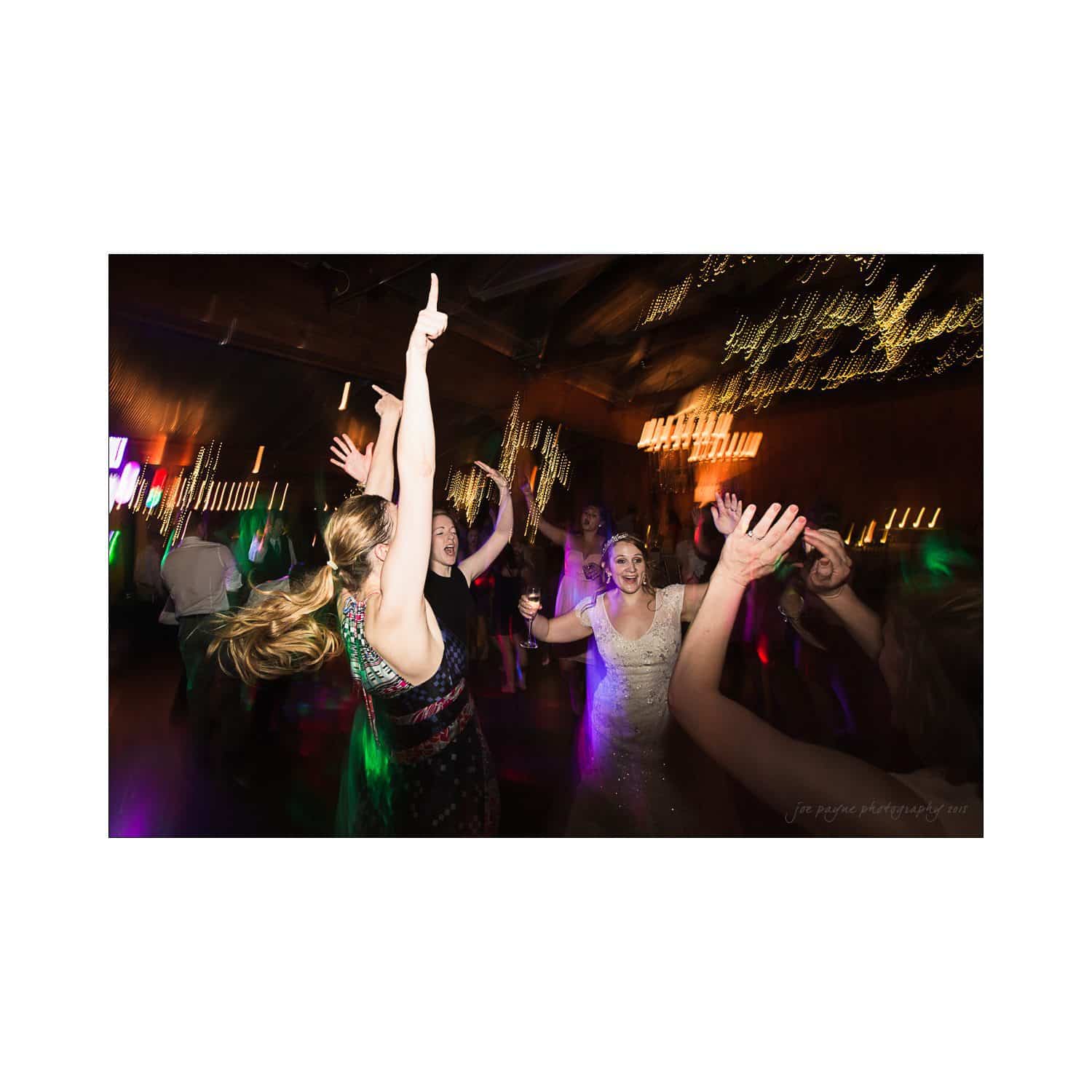 A group of people dancing energetically at a party. A bride in a white dress is joyfully raising her arms. The scene is vibrant with colorful lights and a festive atmosphere.