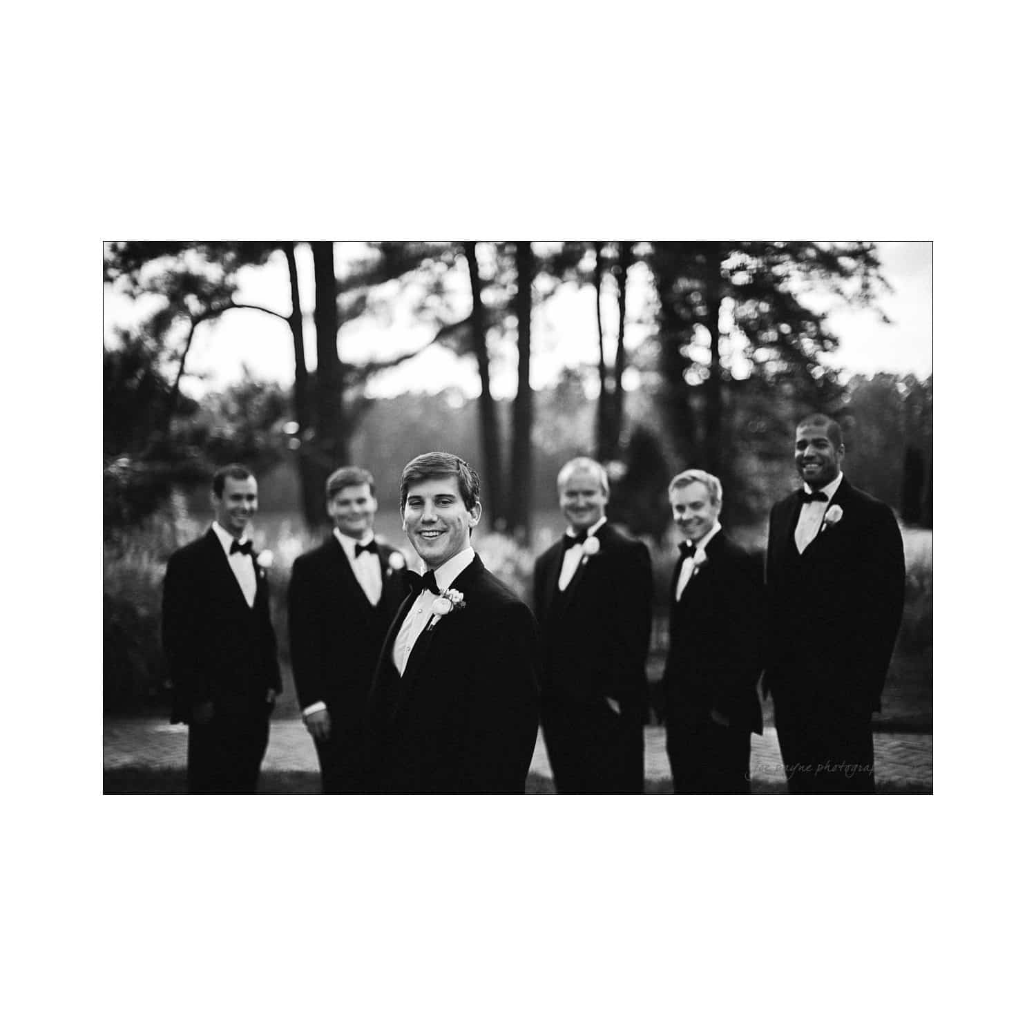 A black-and-white photo of six men in tuxedos standing outdoors on a pathway, surrounded by trees. The man in the foreground smiles at the camera, while the others stand behind him, posing in a relaxed manner.