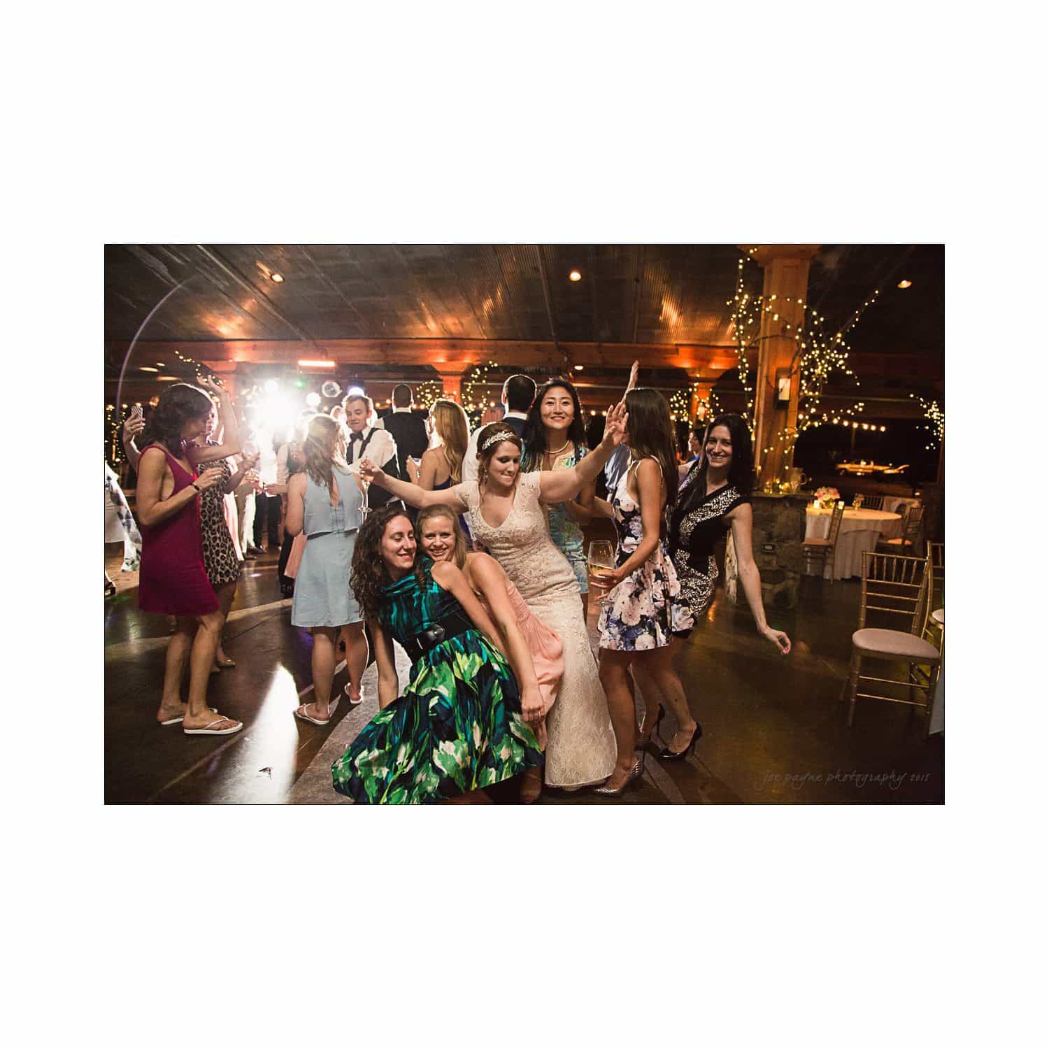 A group of women in colorful dresses, including a bride in a wedding gown, joyfully dance together at a lively indoor reception. Warm lighting and decorative lights create a festive atmosphere.