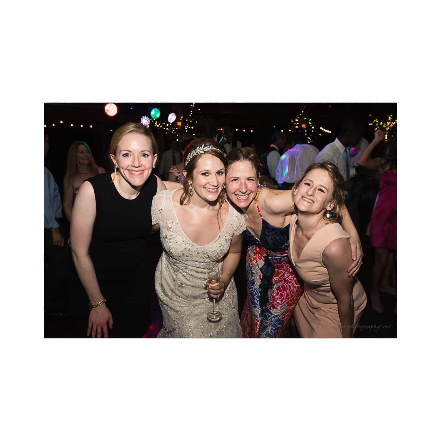 Four women smiling and posing together at a party. The woman in the middle wears a white dress and holds a wine glass. The background shows people dancing and colorful lights.
