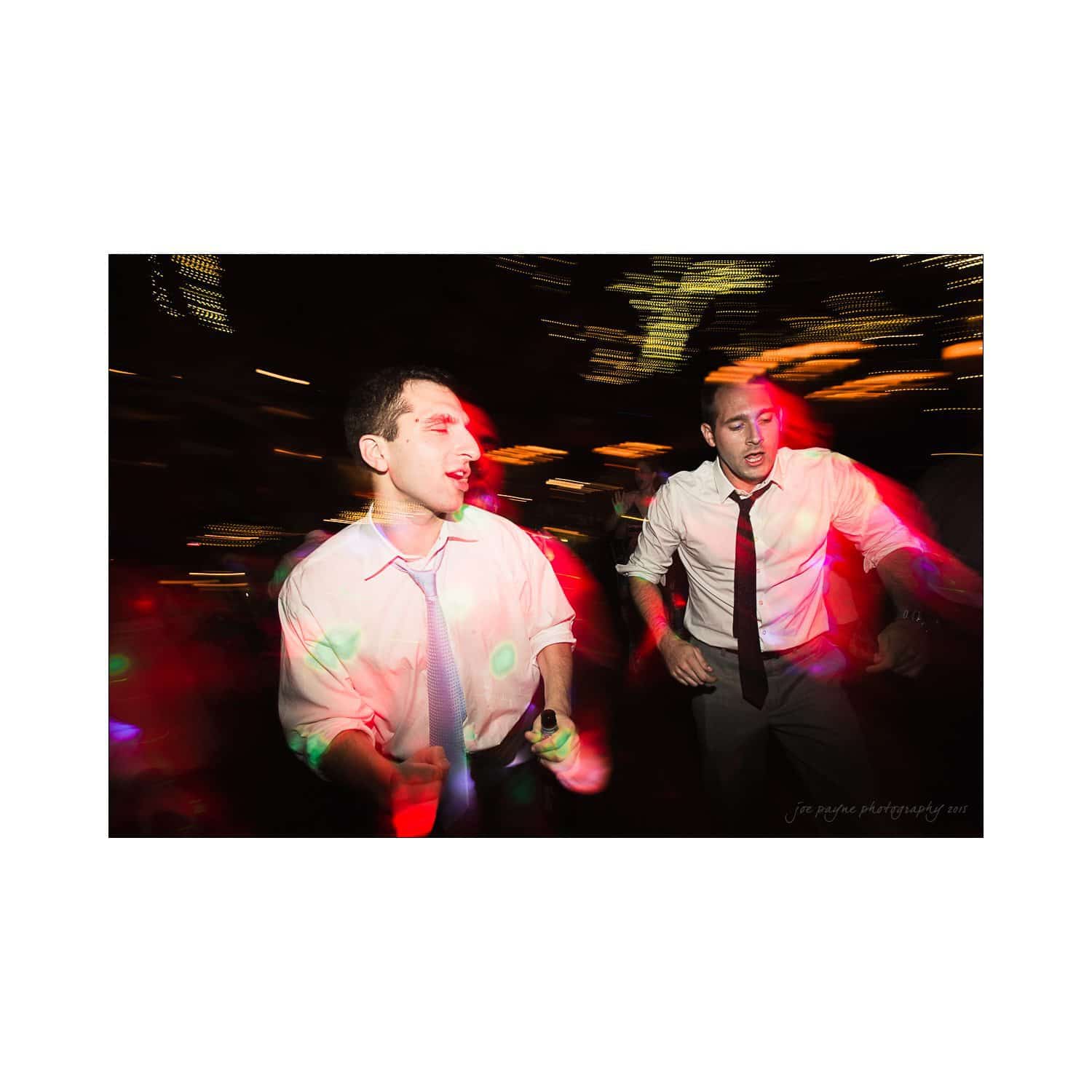 Two men dancing energetically in a dimly lit setting. Both wear white shirts and ties, with colorful lights creating a dynamic atmosphere around them. The image captures movement and a sense of celebration.