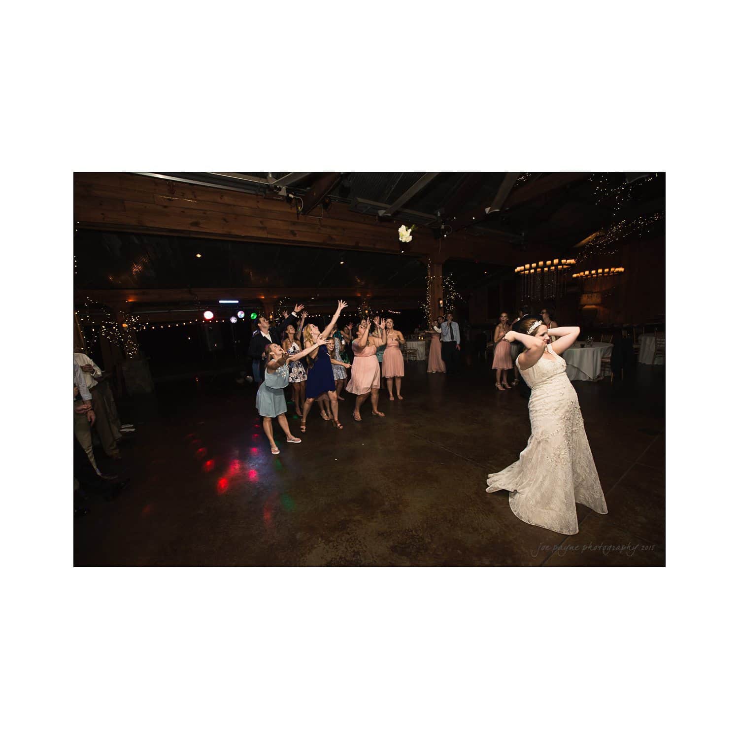 A bride in a white dress is throwing her bouquet to a group of eagerly waiting women in colorful dresses at a dimly lit venue decorated with string lights. The floor is shiny and there are people in the background.