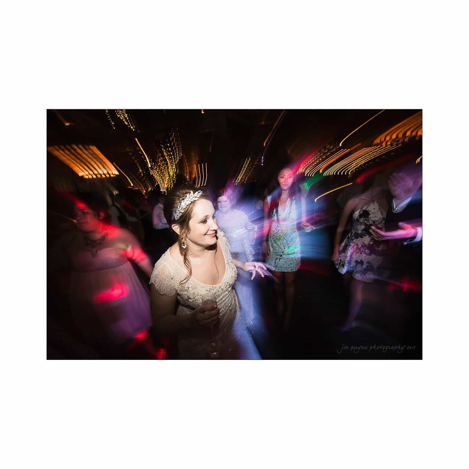 A woman in a white dress dances at a lively party, surrounded by colorful lights and blurred figures, creating a dynamic and energetic atmosphere.