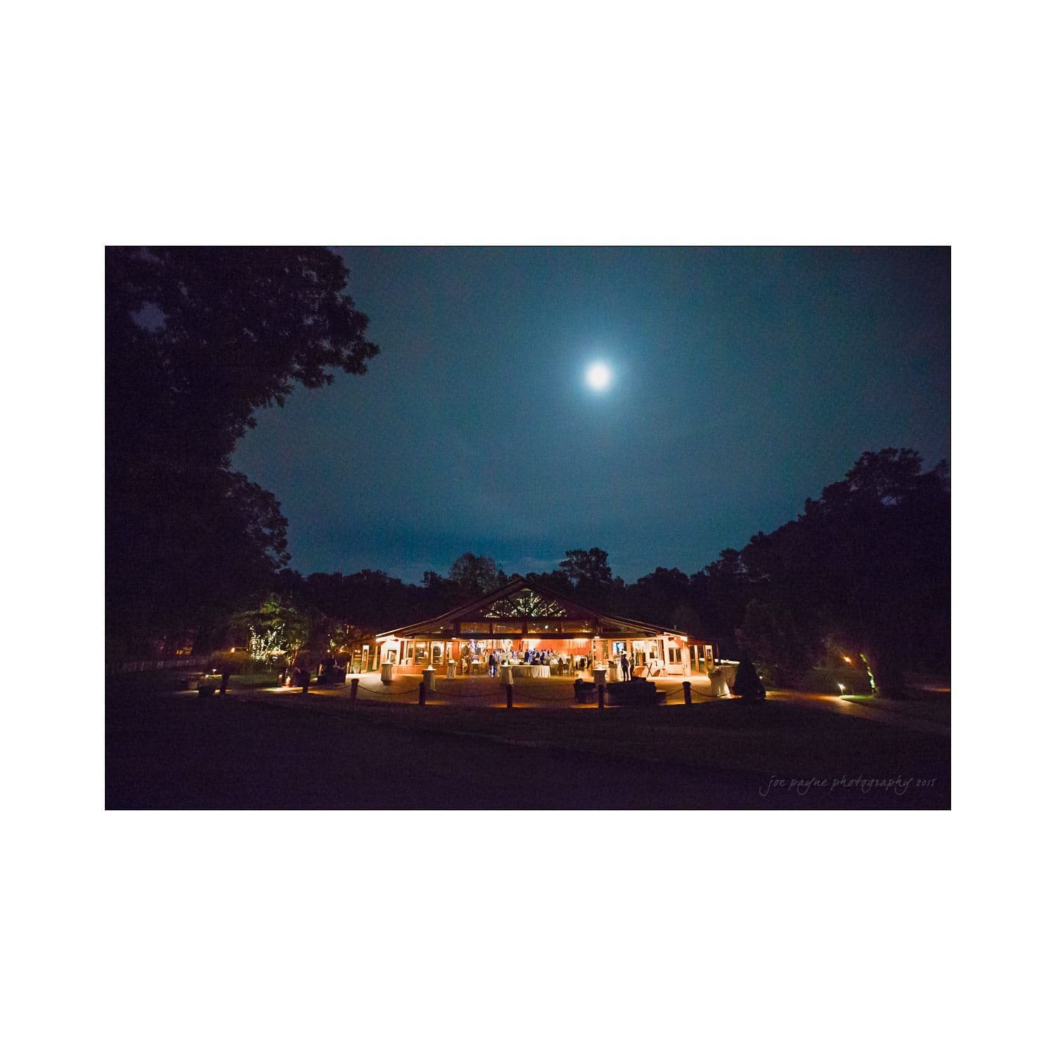 A nighttime scene featuring a well-lit building with string lights, surrounded by trees under a full moon. The sky is dark with a few clouds, creating a peaceful and serene atmosphere.