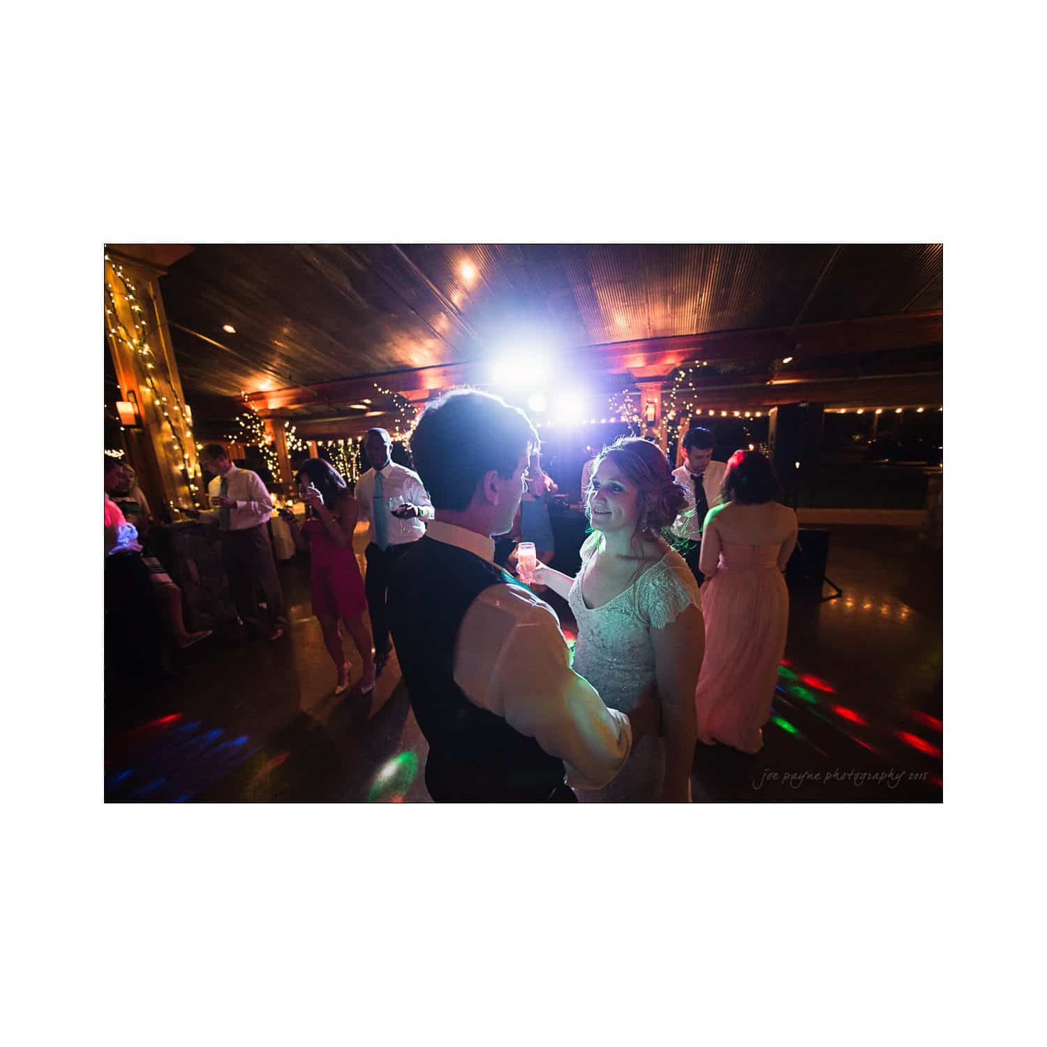 A couple dances closely on a dimly lit dance floor with colorful lights. Other guests are visible in the background, mingling and enjoying the intimate, festive atmosphere.