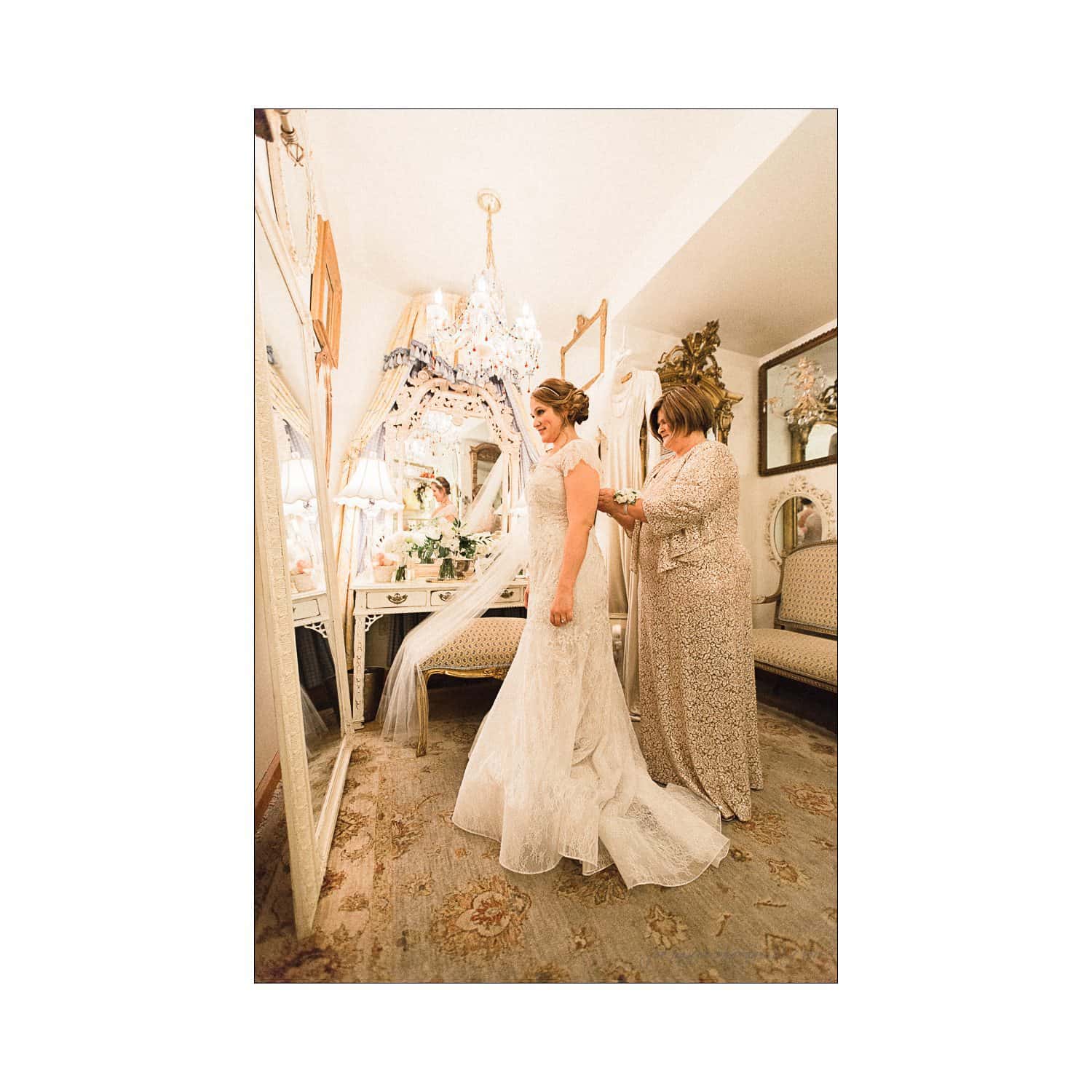 A bride in a white gown stands in an elegantly decorated room, while an older woman in a beige dress helps adjust her dress. The room features a mirror, chandelier, and floral arrangements.